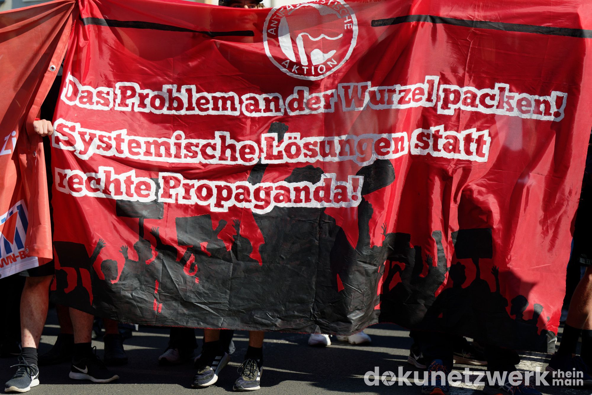 Ein Transparent.Es ist rot schwarz. Oben in der Mitte ist ein Antifa-Logo abgebildet. Darunter steht in roten Buchstaben mit weißen Rand: "Das Problem an der Wurzel packen! Systemische Lösungen statt rechte Propaganda