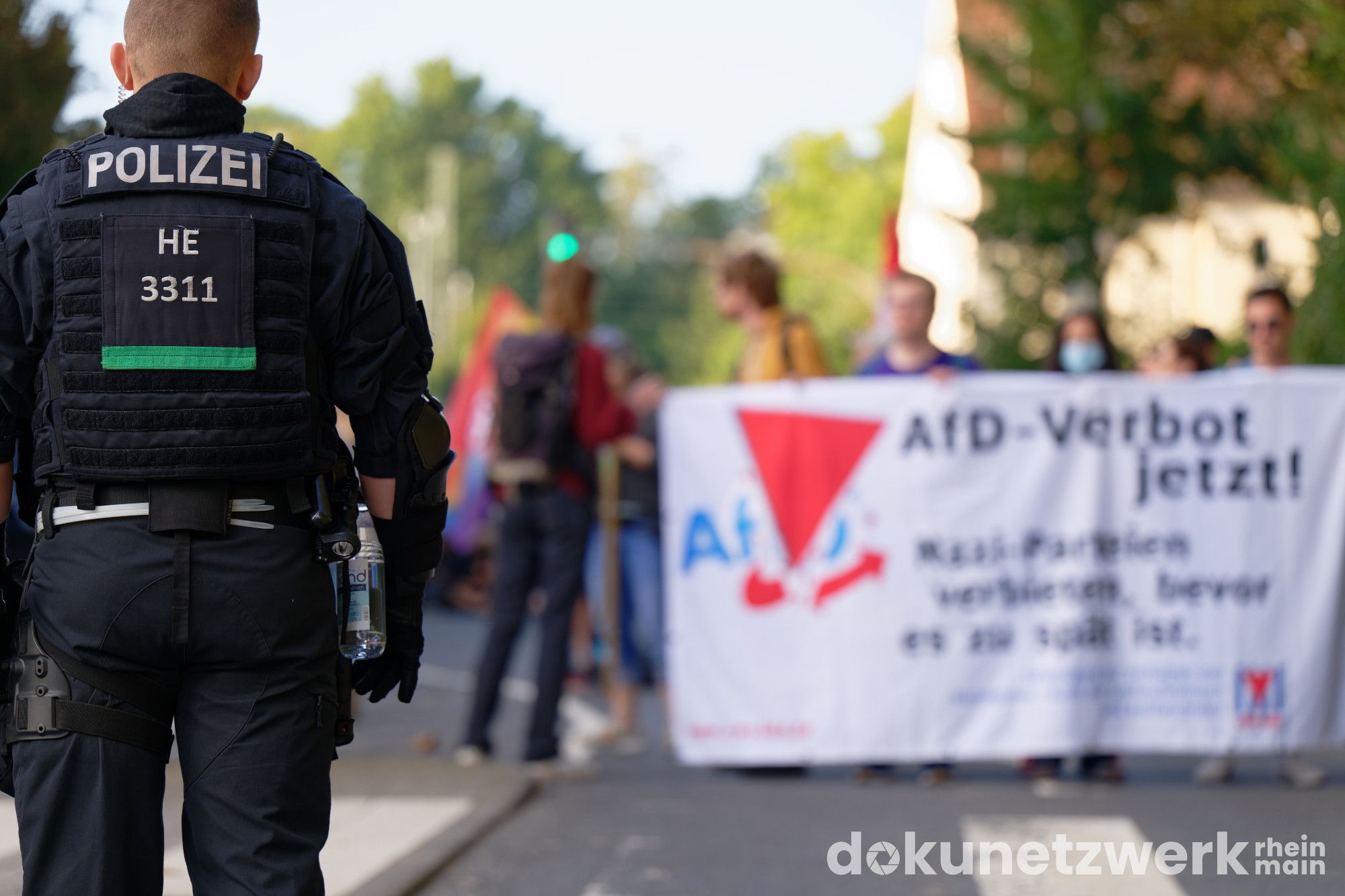 Am linken rand im Vordergrund steht ein Polizist mit dem Rücken zur Betrachtung. Tiefer im Bild sind Personen zu sehen die ein Transparent halten. Darauf ist zu lesen "AfD-Verbot jetzt".