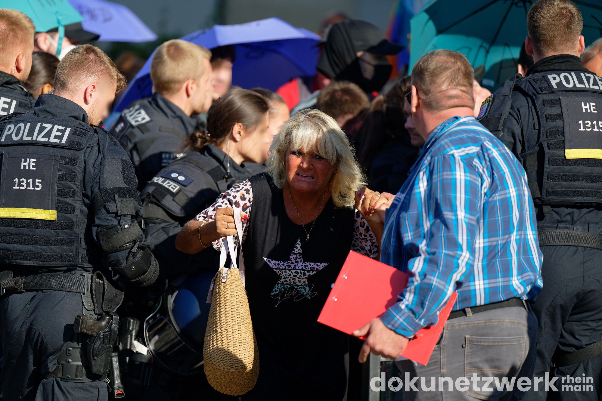 Eine blonde Frau drückt sich sichtlich abgemüht durch die Polizei. Ein Mann mit einer roten Mappe in der Hand hält ihre Hand