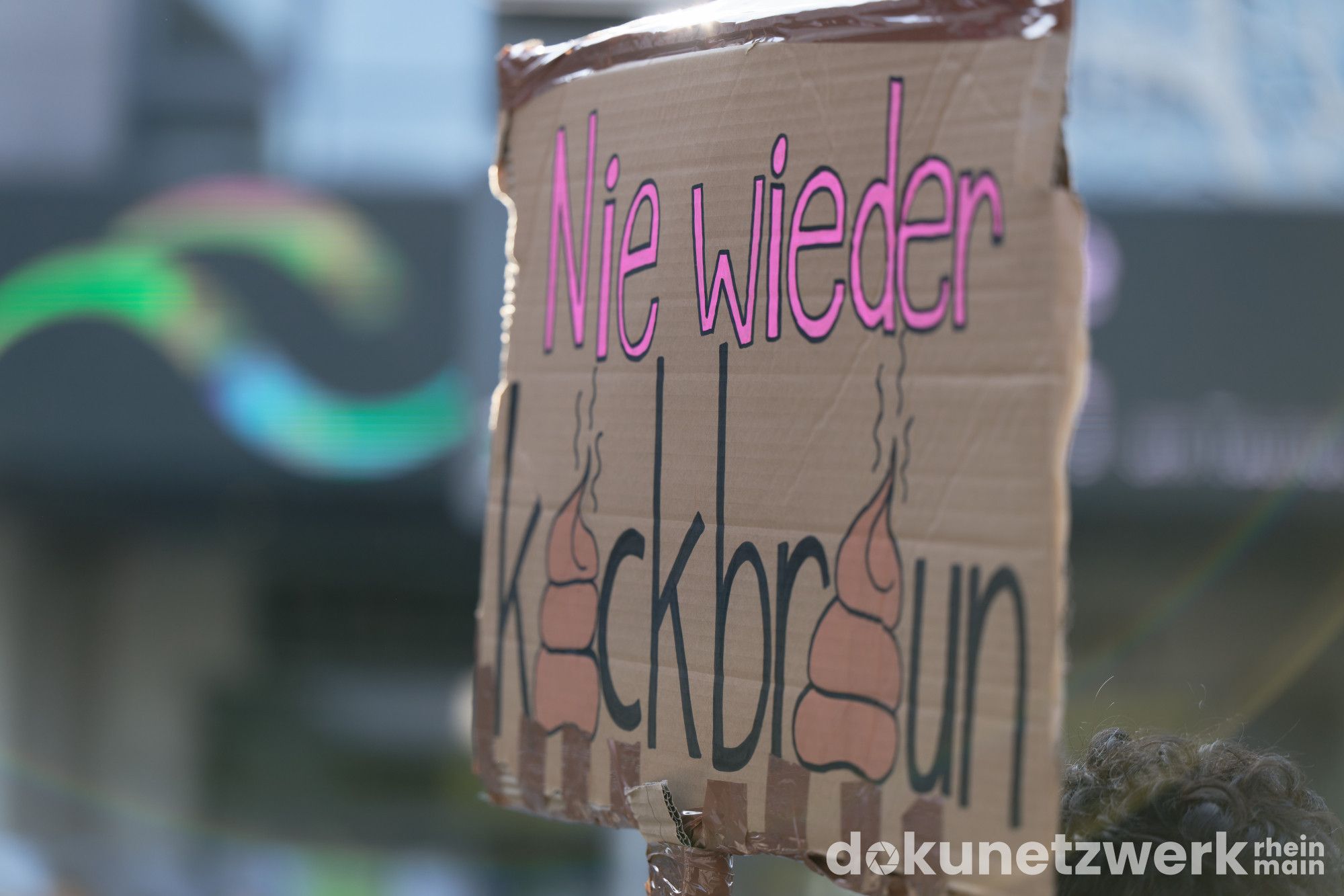 Ein Schild mit der Aufschrift "Nie wieder Kackbraun". Am oberen Rand strahlt das Sonnenlicht drauf. Links daneben sind durch Brechung des Sonnenlichtes bunte Streifen in der Tiefenschärfe