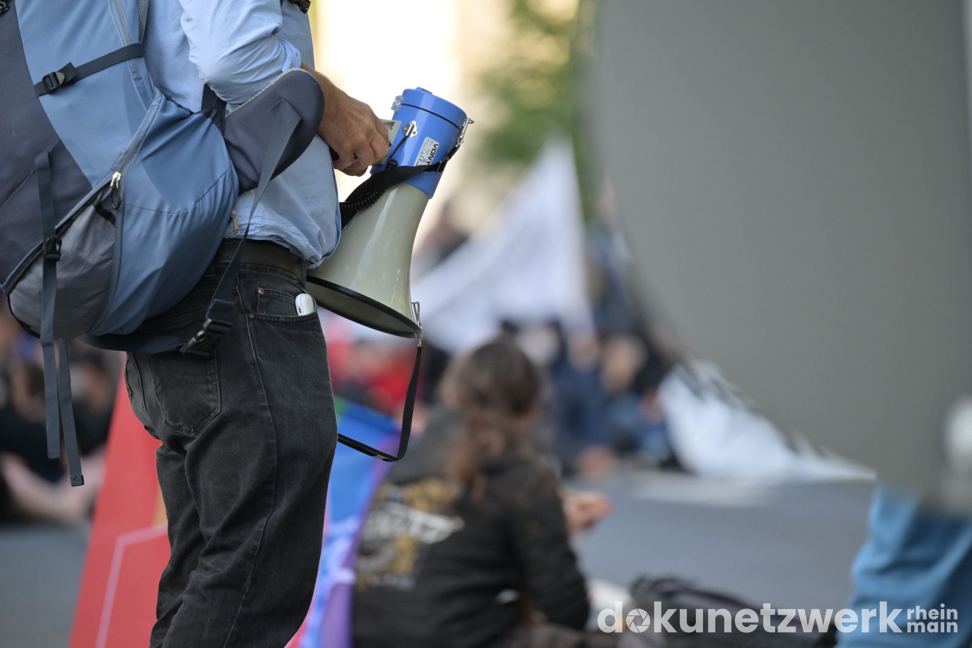 Im Vordergrund ist der Unterkörper eines Menschen zu sehen, welcher ein Megafon hält. Im Hintergrund in der Tiefenschärfe sind schemenhaft sitzende Menschen zu sehen