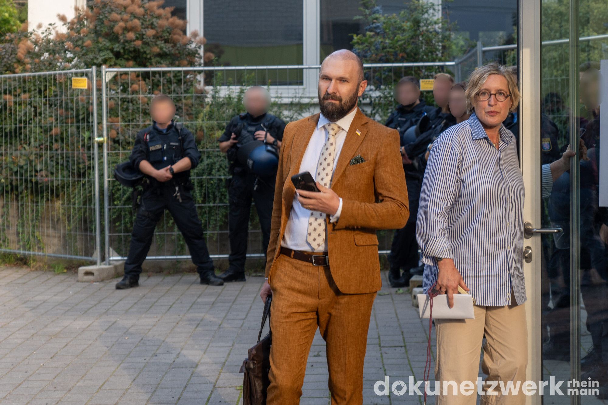 Dimitri Schulz mit Handy in der rechten Hand schaut richtung Kamera. Neben ihn eine Frau, die eine Türe mit Glasscheibe öffnet.