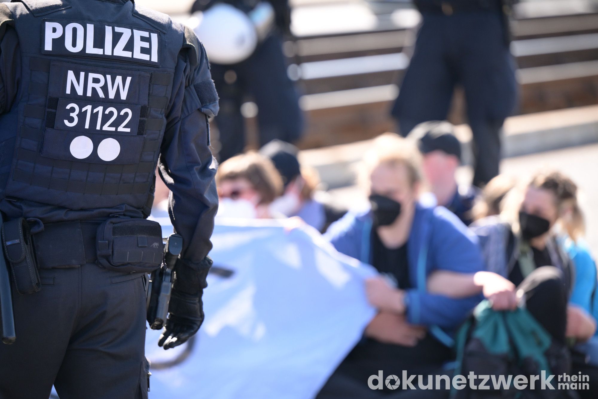 Vorn ist der Rücken eines Polizisten zu sehen. Im Hintergrund sitzen Menschen in einer Sitzblockade