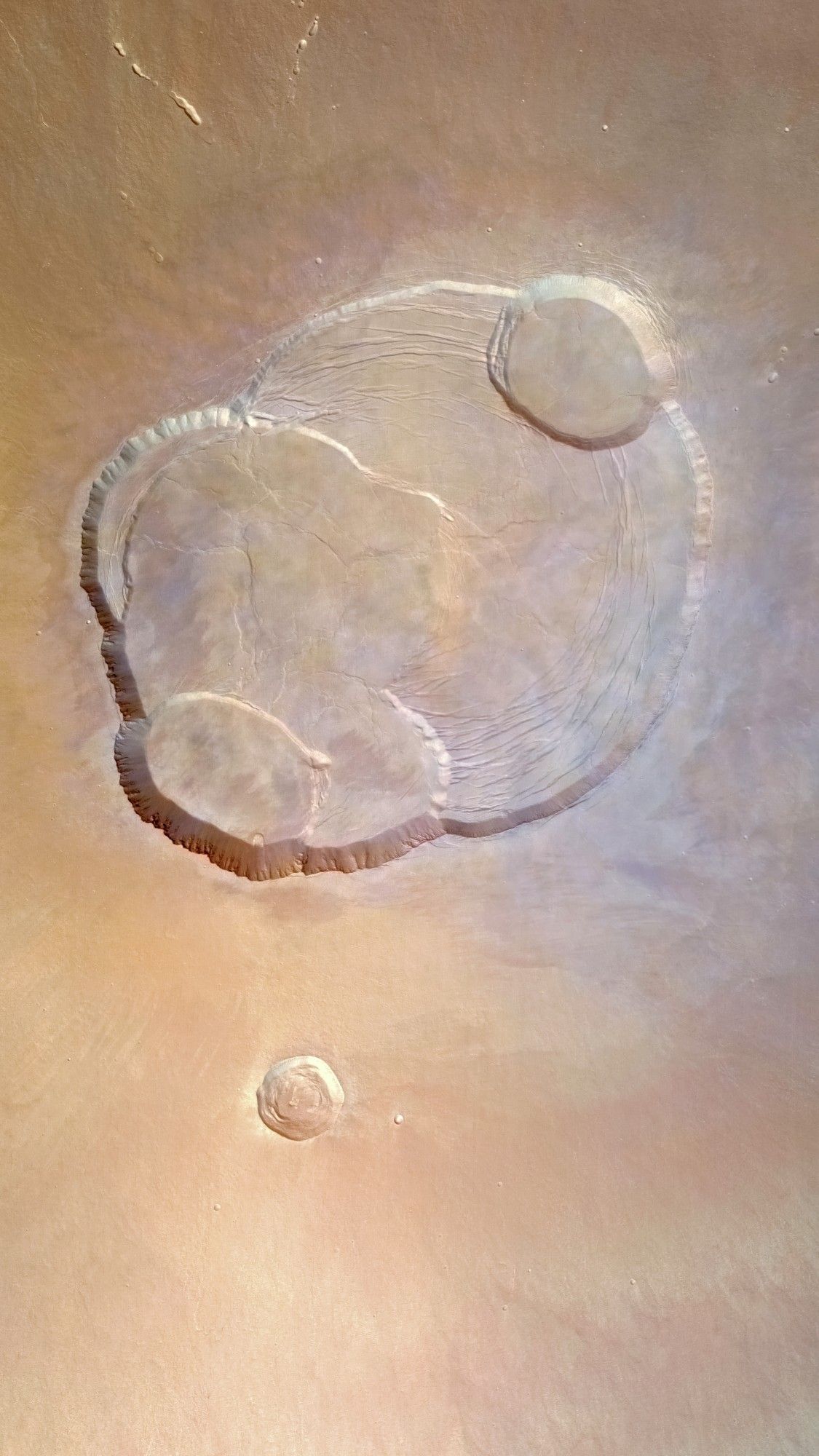 A high-resolution image of Mars' Olympus Mons caldera captured by the ESA's Mars Express. The image shows a large, circular volcanic caldera with several concentric depressions and ridges. The colors are rusty red. Very light clouding is visible on the caldera.