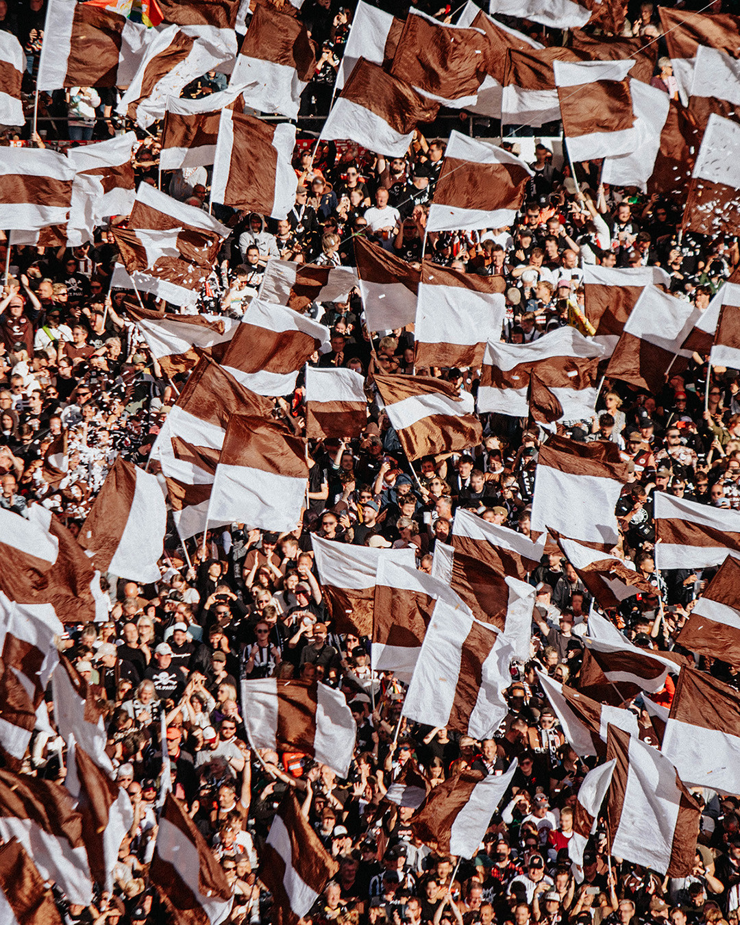 Zahlreiche Fans auf der Tribüne eines Fußballstadions schwenken braun-weiße Fahnen. Die dicht gedrängte Menge erzeugt ein Meer aus wehenden Flaggen.