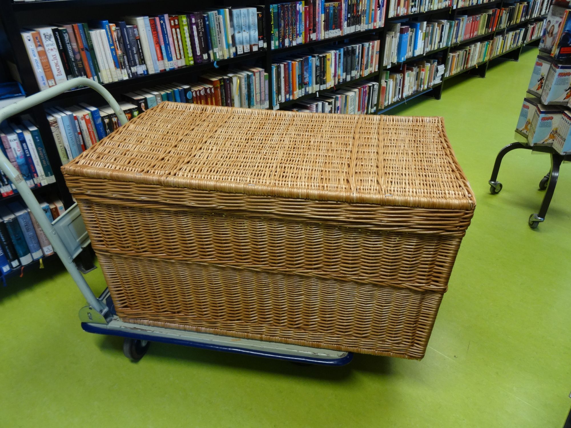 A very big Basket on a trolley in our schoollibrary in front of full bookshelfs, the floor is green (not my favorite color)