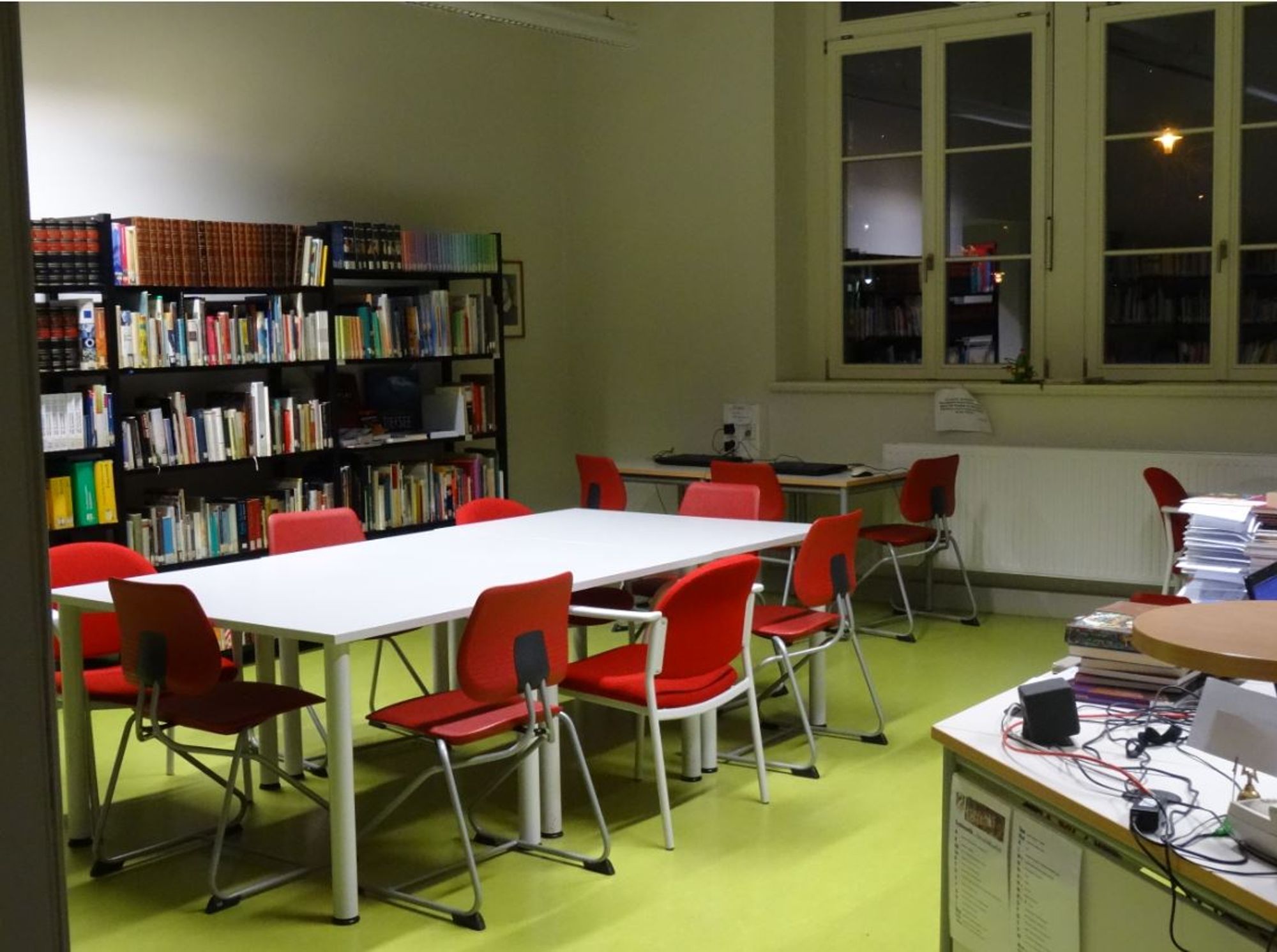 Schoollibrary with white tables and red chairs, shelfs with books at the left side, windows in the background, dark outside, at the right side my working place