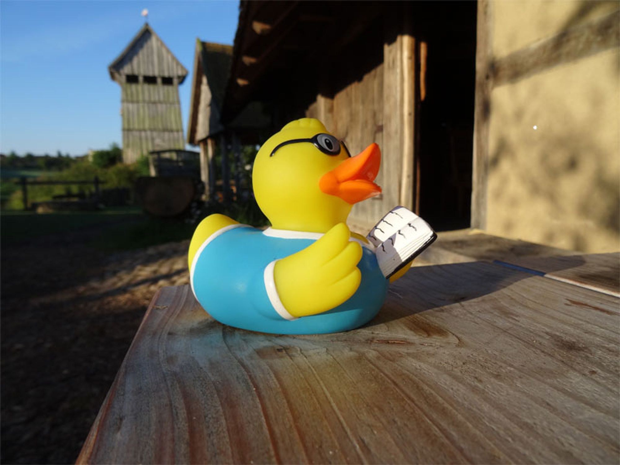 A reading rubber duck on a wooden table in front of a early medieval house, in background a wooden tower