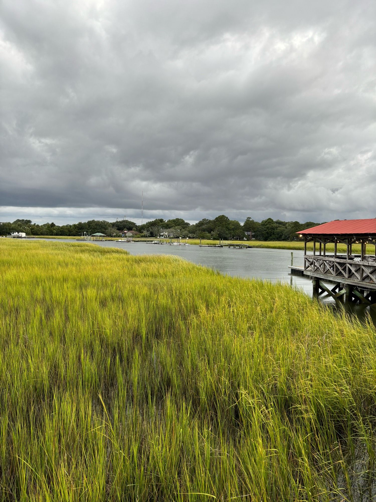 View of a waterway and pier. The sky is overcast and a little threatening. Boats can be seen anchored in the distance. The scene is peaceful and quiet.