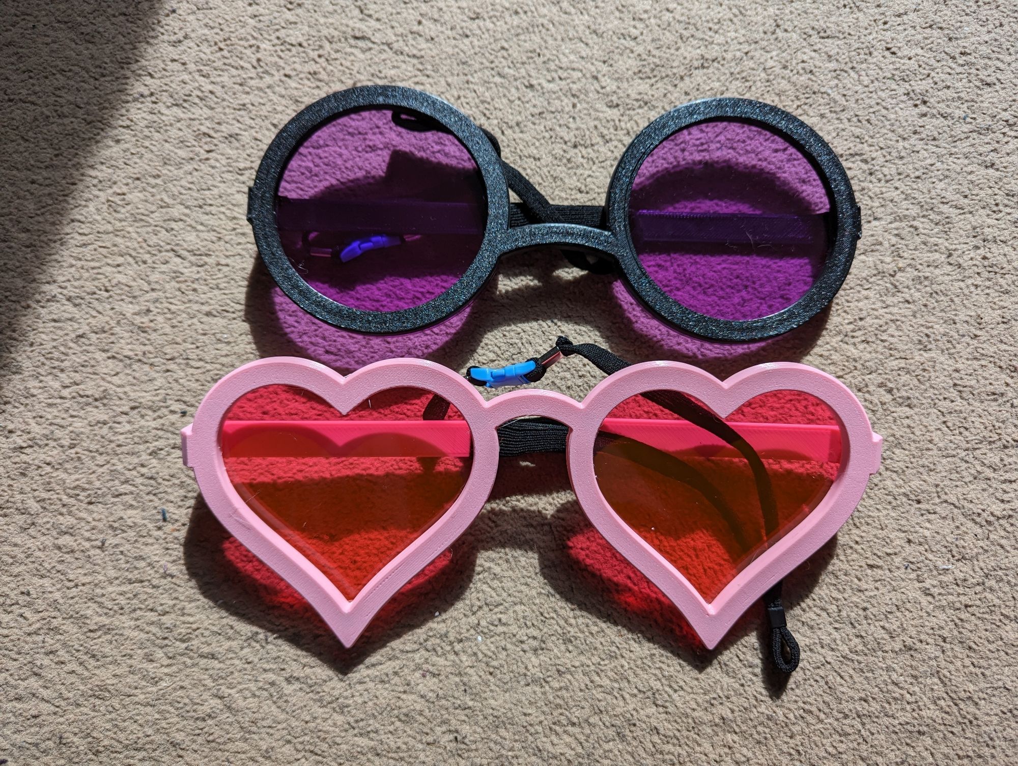 A pair of black circular glasses frames, and pink heart shaped frames, with purple circle and red heart lenses attached magnetically