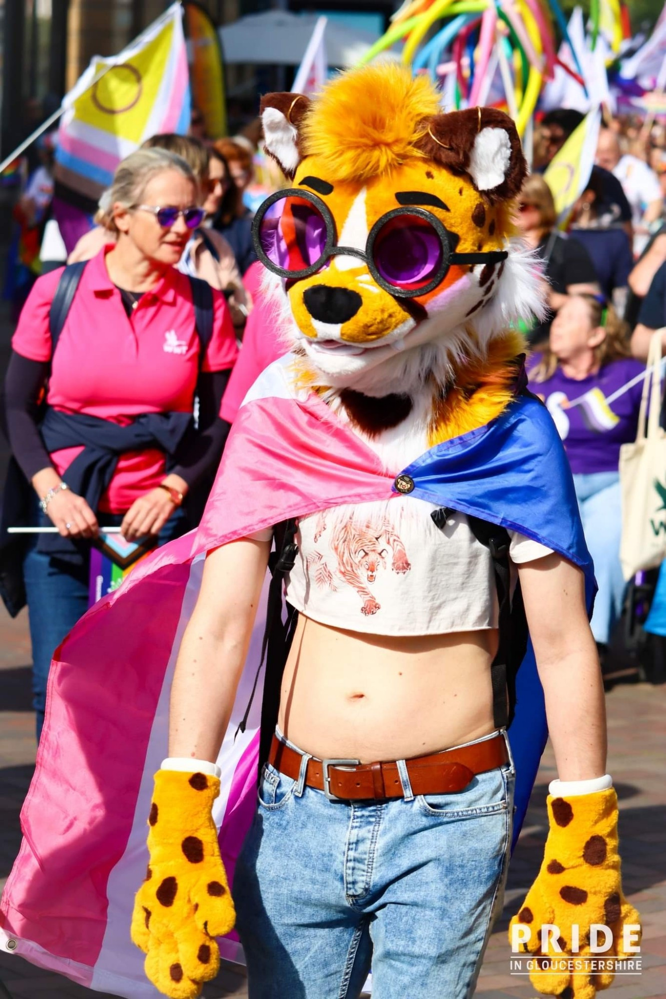 Me at the Gloucestershire Pride parade on 15 September. Wearing my cheetah partial fursuit, crop top, and genderfluid flag as a cape. A bunch of other queers are in the background