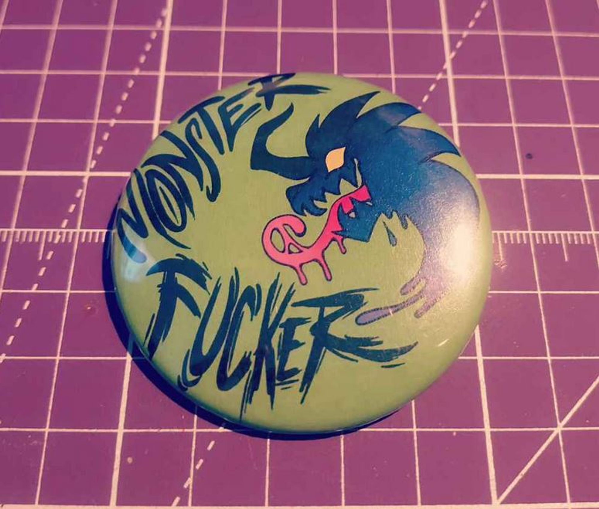 A lime green "Monster Fucker" button featuring a silhouette of a behemoth-like monster with a long pink drooly tongue, horns, and yellow eyes.