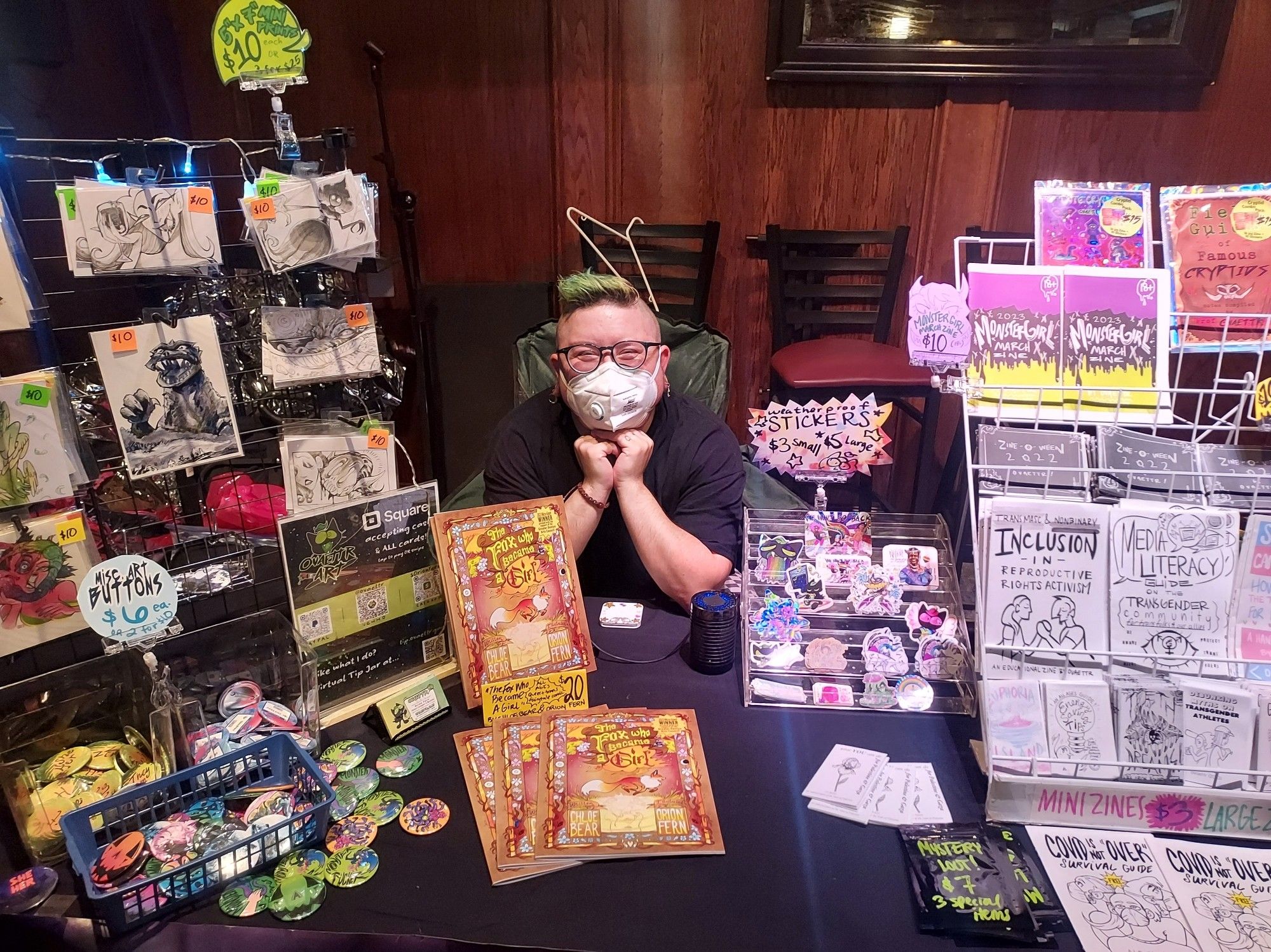 Me sitting behind my display table featuring zines, comics, stickers, buttons and prints.