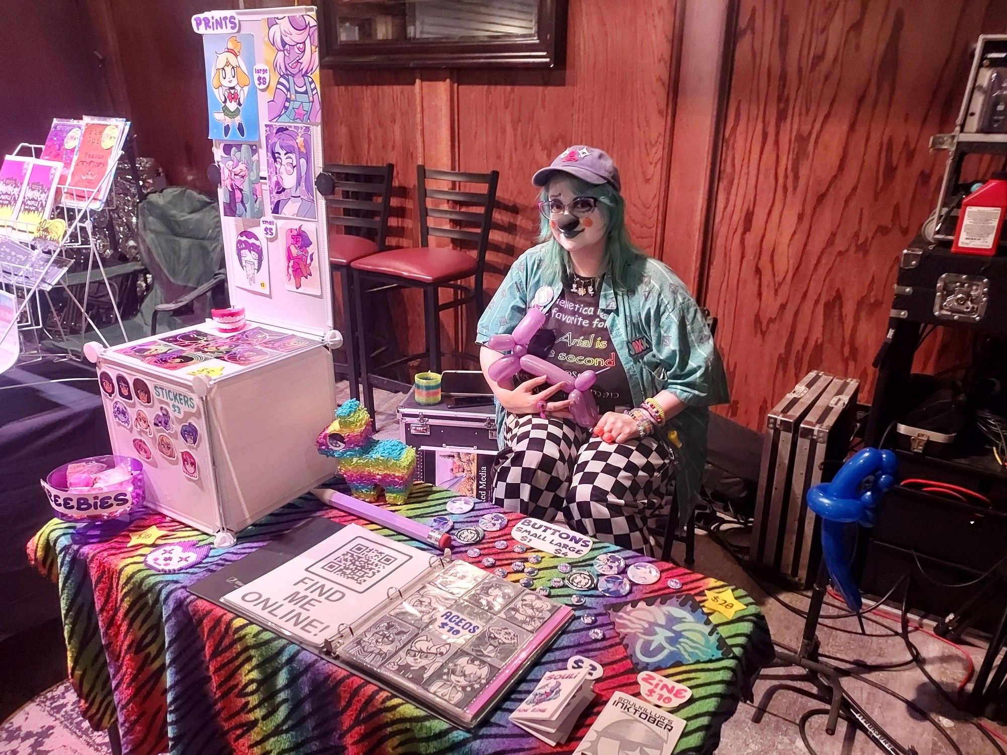 A cool clown guy with a black clown nose , baja blast hair and a neat funky outfit with patterns galore. He is sitting behind a vendor table.