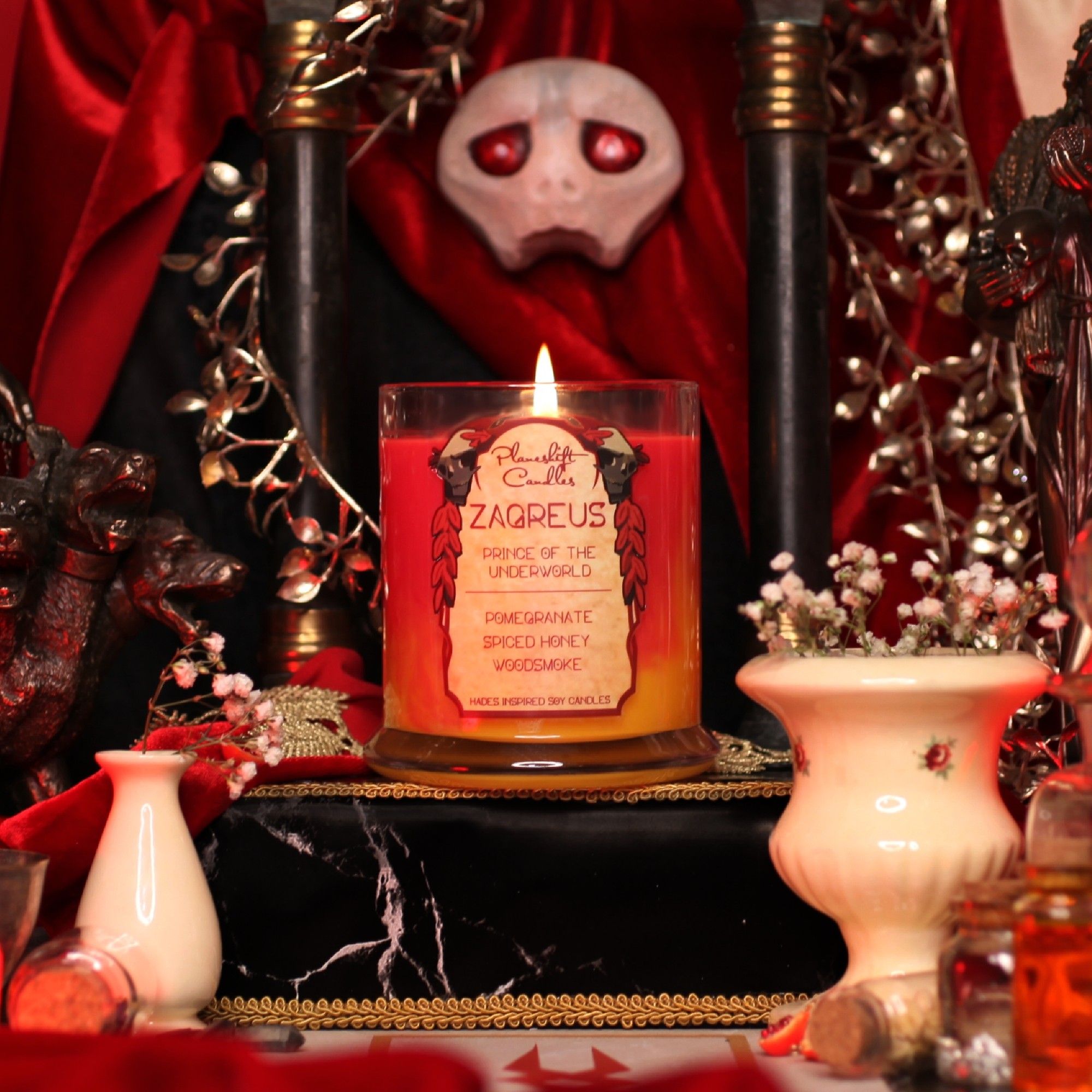 A red and yellow/orange coloured candle with a label that reads "Planeshift Candles - Zagreus, Prince of the Underworld" listing pomegranate, spiced honey and woodsmoke as the fragrance.