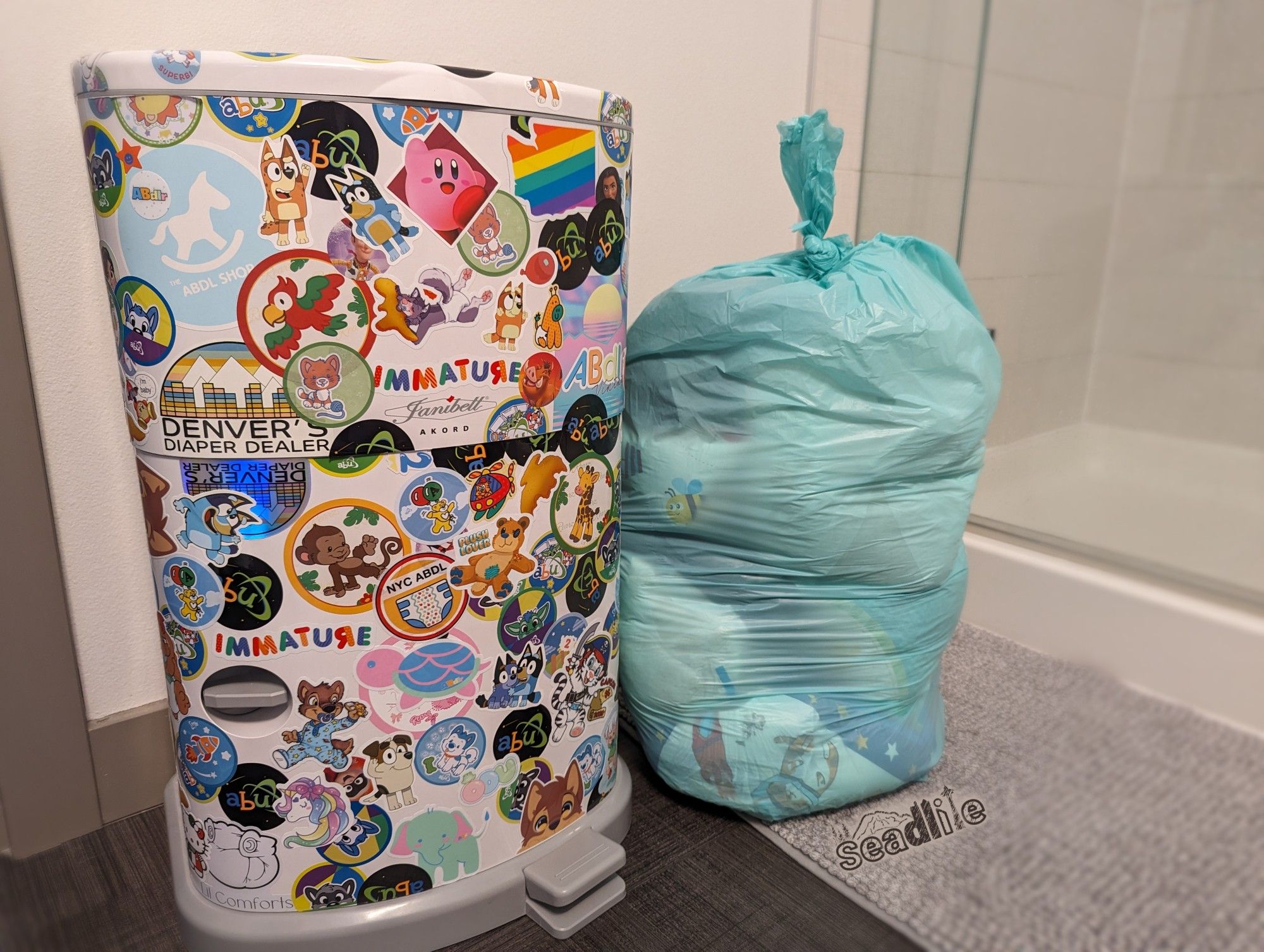 A Janibell Akord 330 diaper pail colored in colorful ABDL, babyfur, and cartoon stickers. Next to the pail on the floor is a teal plastic pail bag full of soggy diapers.