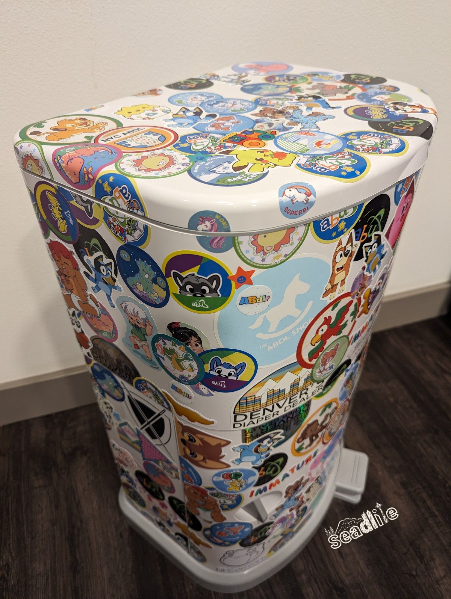 A Janibell Akord 330 diaper pail colored in colorful ABDL, babyfur, and cartoon stickers (left side perspective).