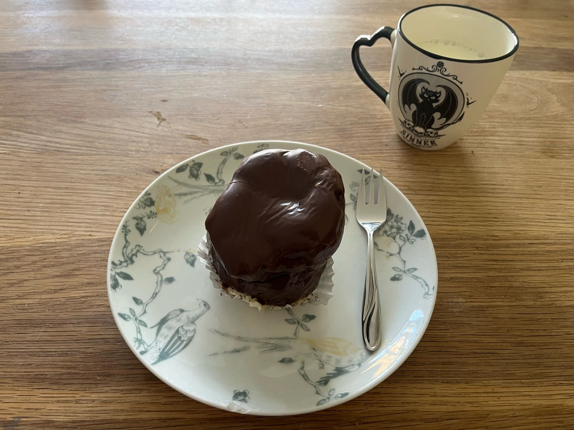 Tafel met daarop een Bossche bol op een bordje, een vorkje en een mok melk met daarop een zwarte kat met vleermuis-vleugels en het bijschrift ‘sinner’.