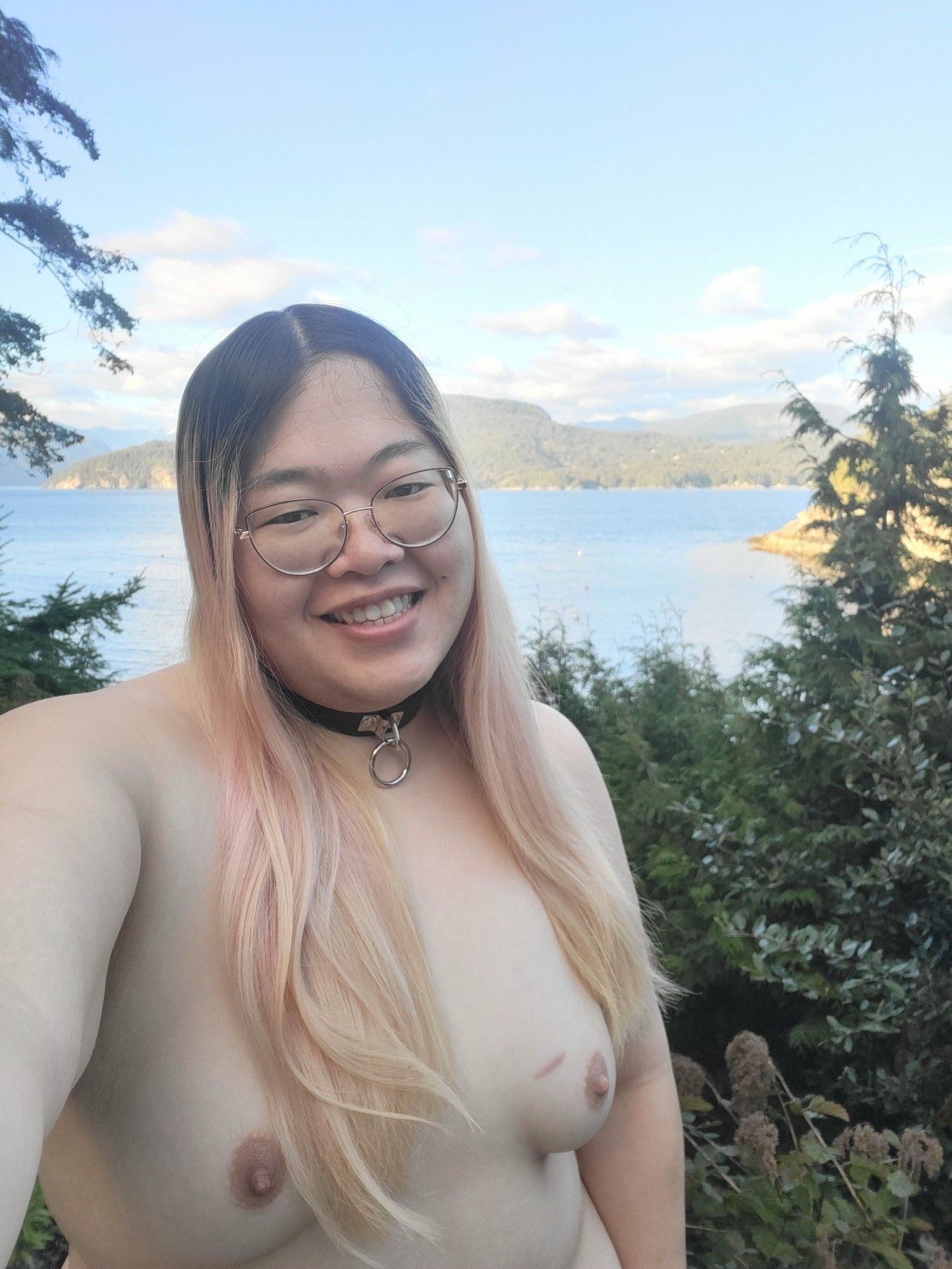 Natalie is standing naked on the patio in front of the waters at a lil cove on the island, in the front of the background is trees ans greenery, and a bit further is a pretty view of the water and the island behind her