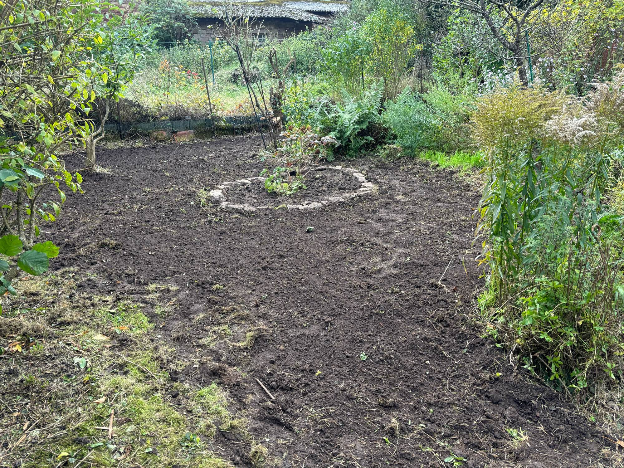 Umgegrabene Fläche, darin ein rundes Beet mit Natursteinen umrahmt, im Hintegrund Zaun