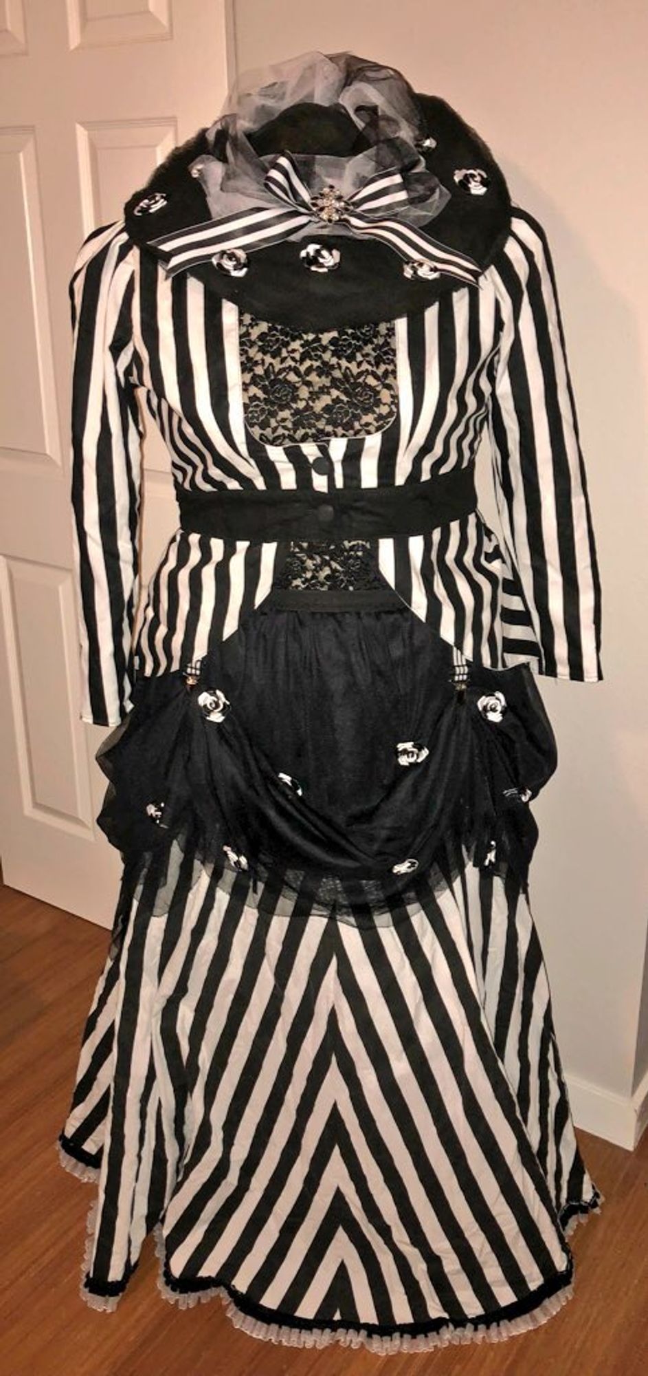 An outfit on a dress form: Black & white striped jacket and matching skirt, black tulle overskirt trimmed with black & white ribbon roses, and a broad-brimmed black tulle hat trimmed with black & white tulle, black & white striped bow, and more black & white ribbon roses.