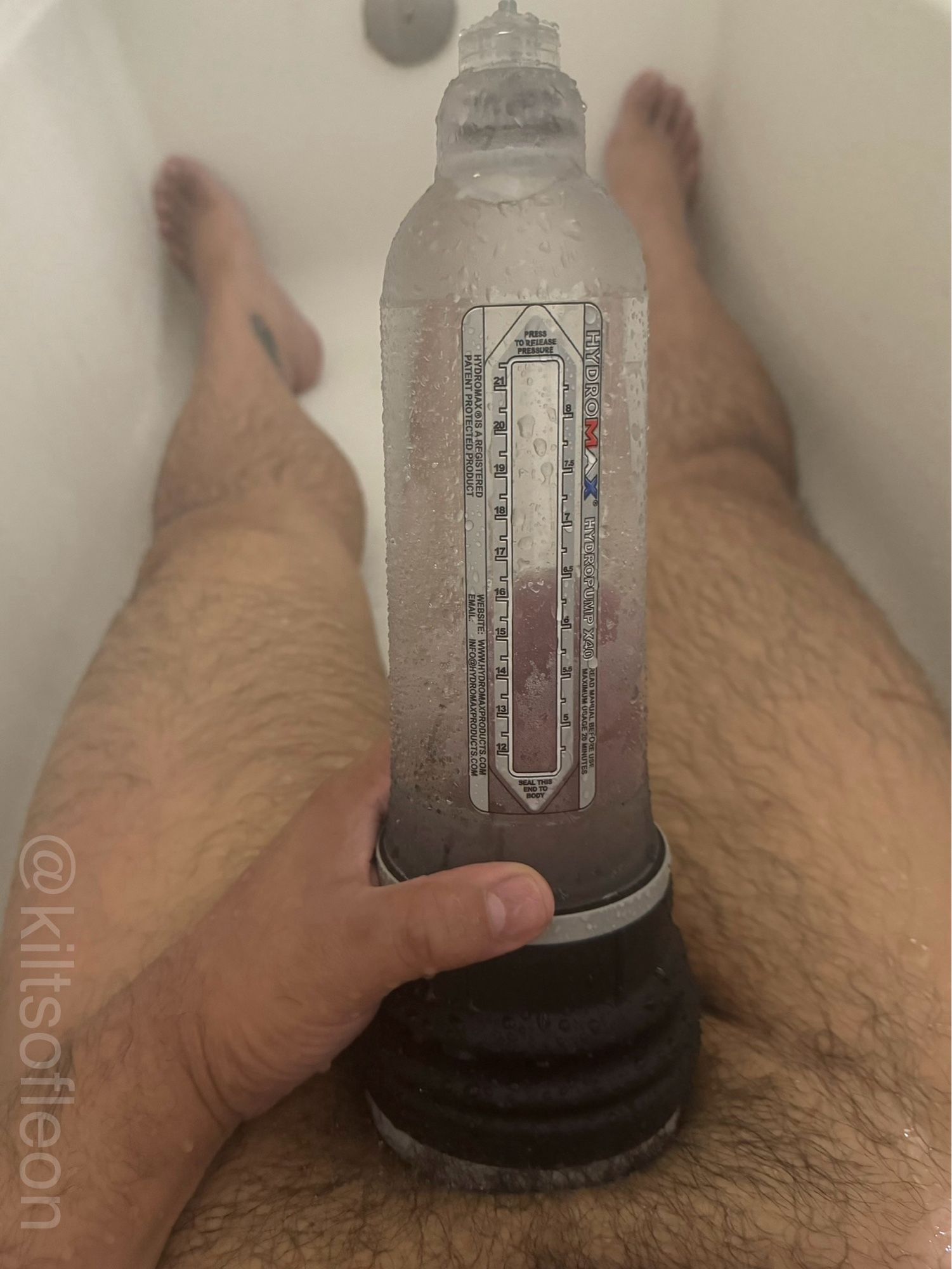 Leon naked sitting in a bathtub, holding a cylindrical pump with a black accordion base against his crotch. The pump is filled with water and contains his dick.