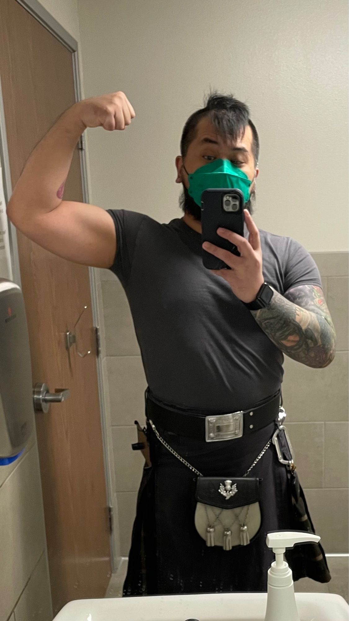 Leon in a laboratory restroom in a plain tshirt, black kilt with brown tartan box pleats, belt with large rectangular buckle, black and white semi dress sporran, and KN95 mask. He’s flexing his left biceps in the mirror because this restroom lighting is flattering.