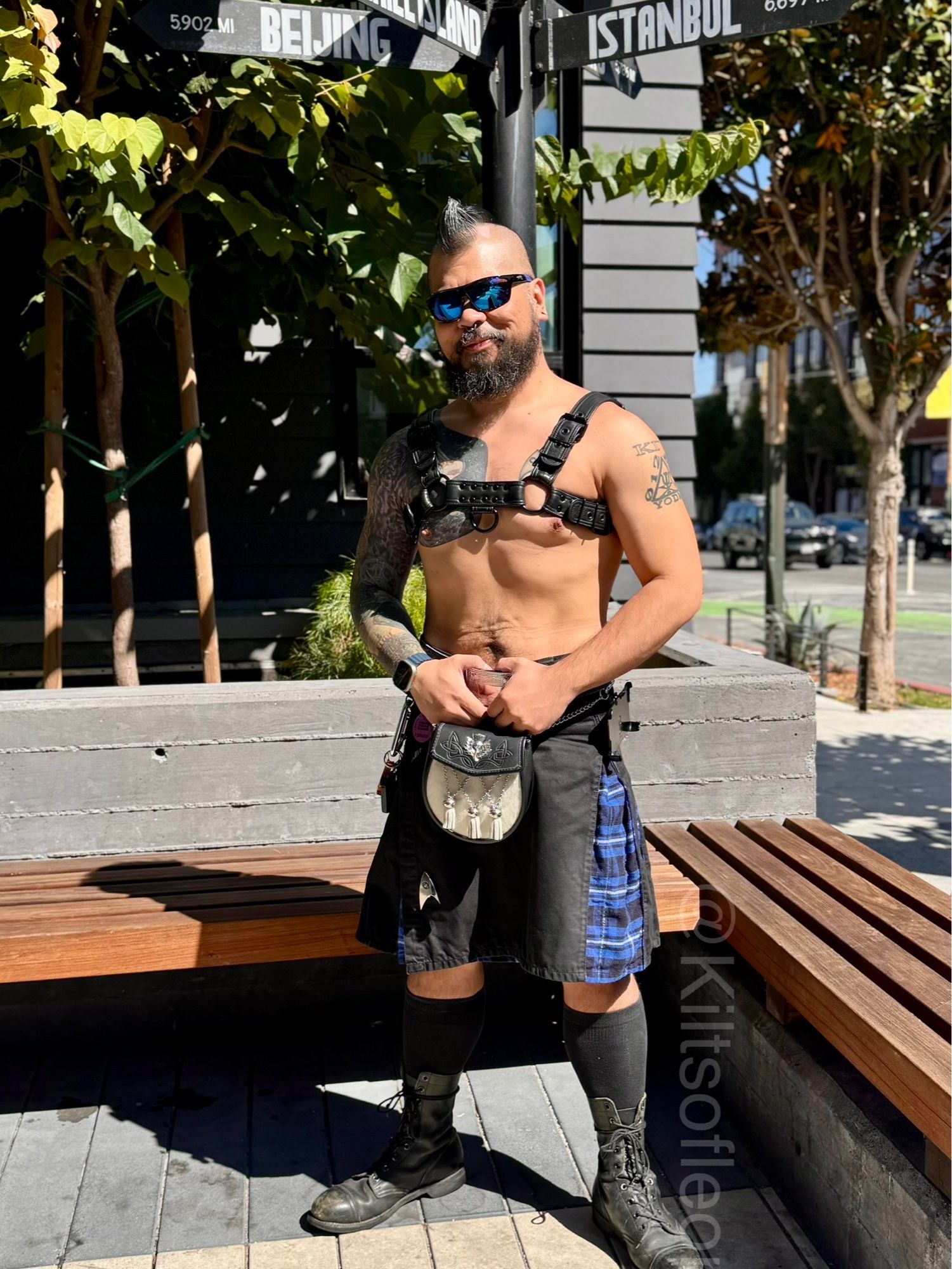 Leon, a mohawked, tattooed man wearing a bulldog harness, combat boots, calf socks, a black kilt with blue, black, and white tartan pleats, and a black and white semi-dress sporran.