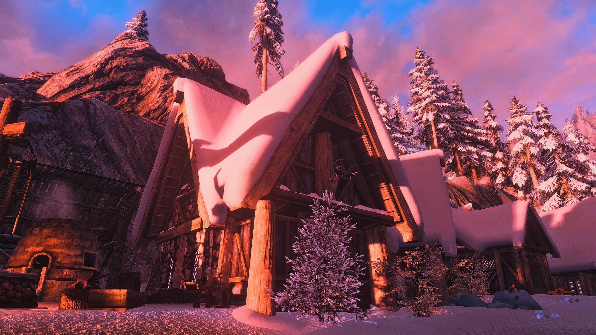 A snowy village at sunrise. Powdery snow covers the ground while piles of fluffy snow coat the roof of each log cabin. The tall trees have snow on their branches as well. A blacksmith forge area is nestled beside the most prominent cabin, with stone from the mountain behind it. The sunrise colors the sky in tones of pink and blue. This is a screenshot of Skaal Village from the Dragonborn DLC in Skyrim Special Edition.