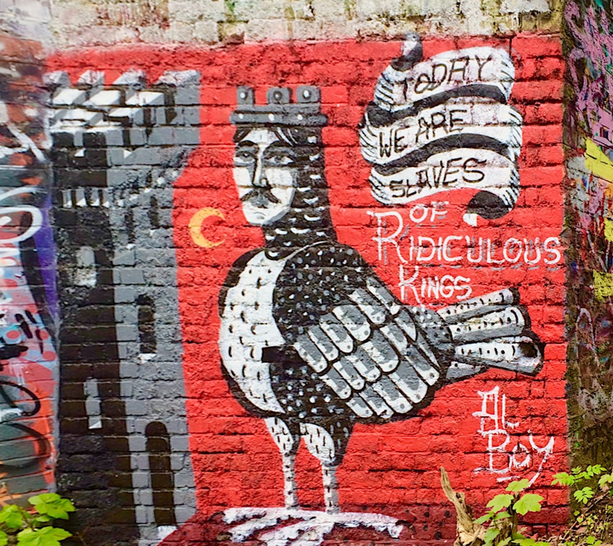 Graffiti painted on brick wall and signed by "Al Boy".

On a red background, a mustachioed and handsome young "king" is drawn, in monochrome grey, as a bird with a man's face. The wings, chest, neck and other feathers are rendered beautifully in heraldic-style patterns. The king wears a crown so you know he's a king. A stylised castle, also purely grey, stands to the left of the bird-king. Elegant ribbons in the upper right read "TODAY / WE ARE / SLAVES" and under this, white lettering reads "OF RIDICULOUS KINGS". A yellow crescent has been placed between the King and his Castle. The red/yellow crescent combination is likely a reference to Islam and maybe even the Crusades, but beyond that, I can't interpret the symbology.