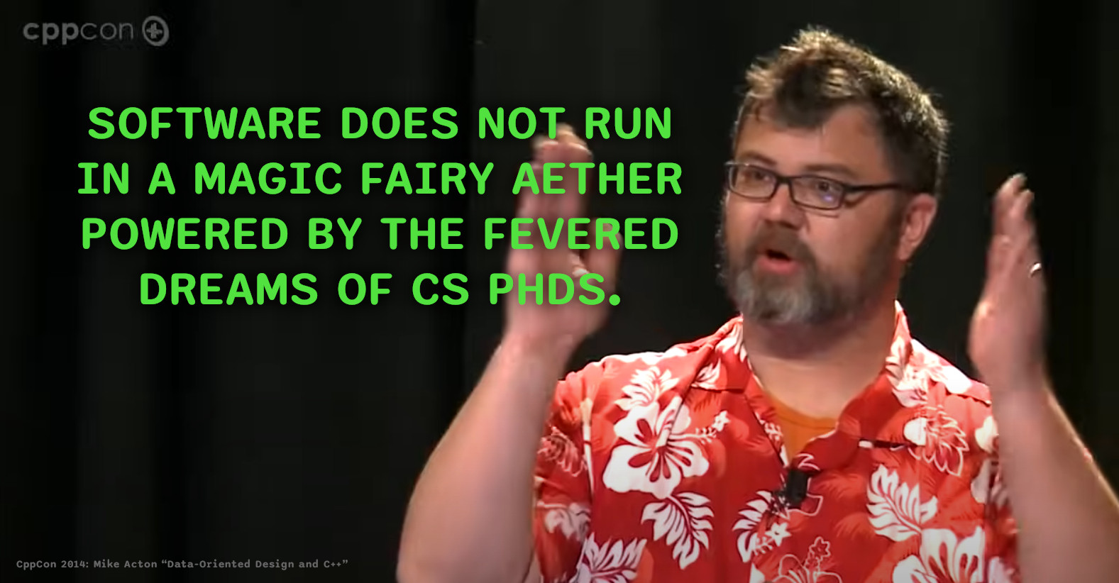 Mike Action making a favourite point in his keynote(?) at CppCon 201: "Data-Oriented Design and C++".

He is in a rather good Hawaiian shirt, a dark orange with white print pattern; he is wearing practical rectangular nerdy glasses; he is bearded with "young dad" level hair; kind of bear-shaped, and he's sweating under the lights and his frenetic movement during the talk. He's brightly lit in front of very dark curtains.

The quote from a slide about to be shown, reads: "Software does not run in a magic fairy aether powered by the fevered dreams of CS PhDs."

Video: https://youtu.be/rX0ItVEVjHc?t=975