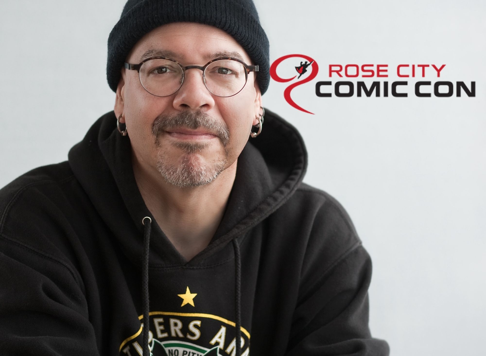 A very out of date head shot of author Greg Rucka wearing a black watchcap and a black Timbers Army sweatshirt with the Rose City Comic Con banner to one side. He seems mild mannered enough.