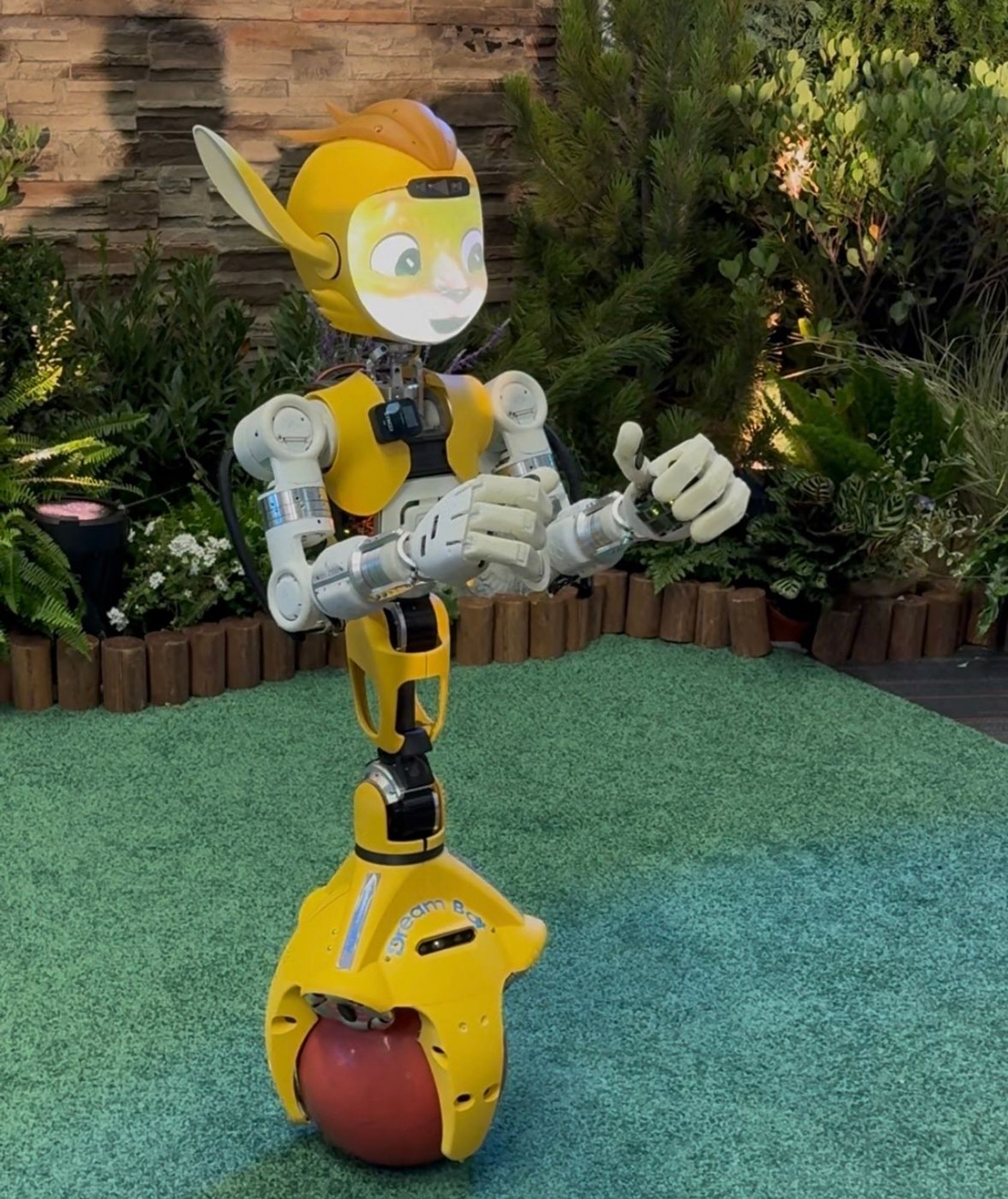 A yellow robot balances itself on a single sphere allowing it to move in all directions easily. The face is digital, appearing as perhaps a cheetah. It has large human like hands.