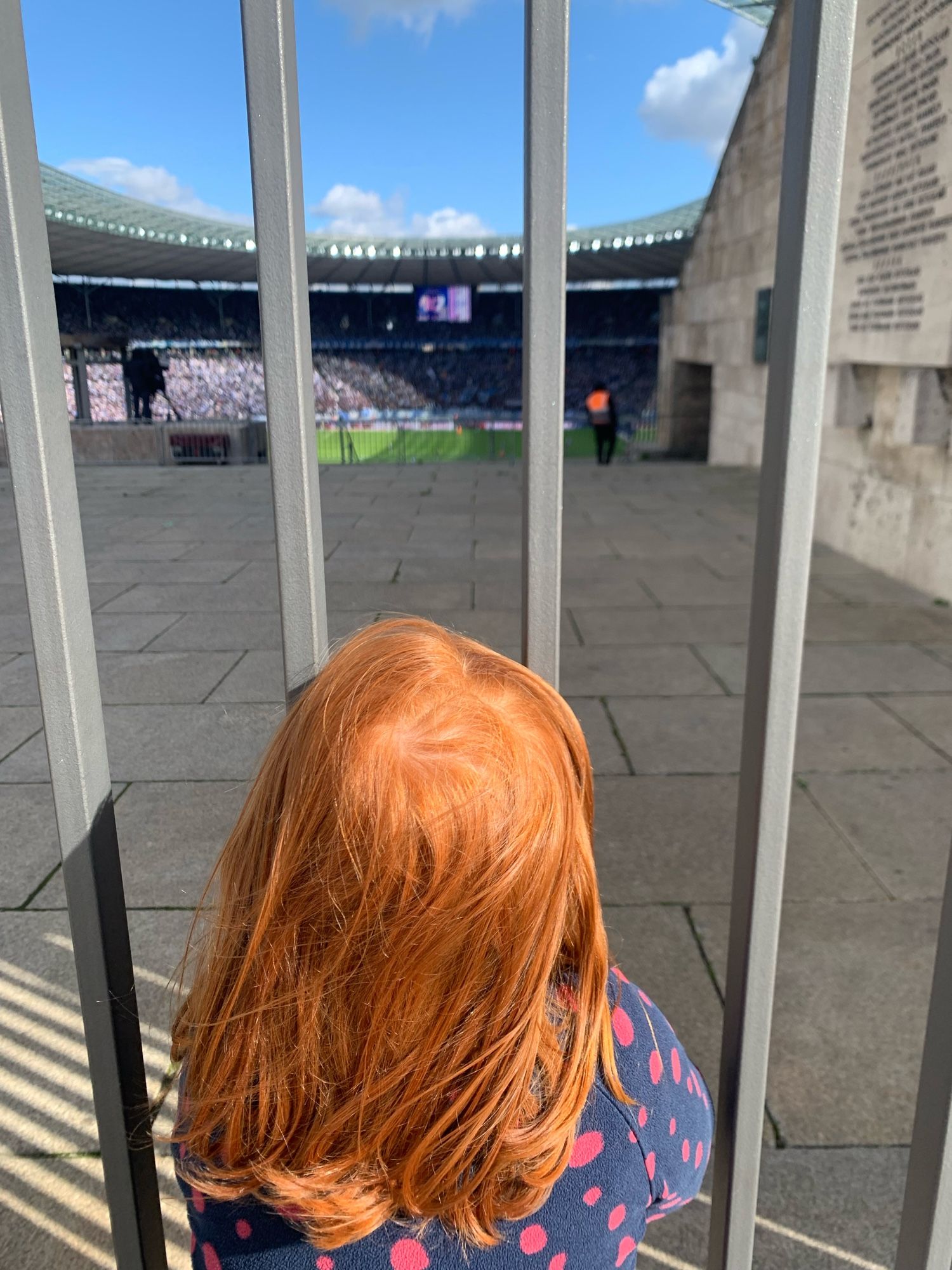 A redhead shown from behind watching a football match through the gates of the Marathontor at the Berlin Olympic Stadium. The sun is shining and all seats in view are filled.