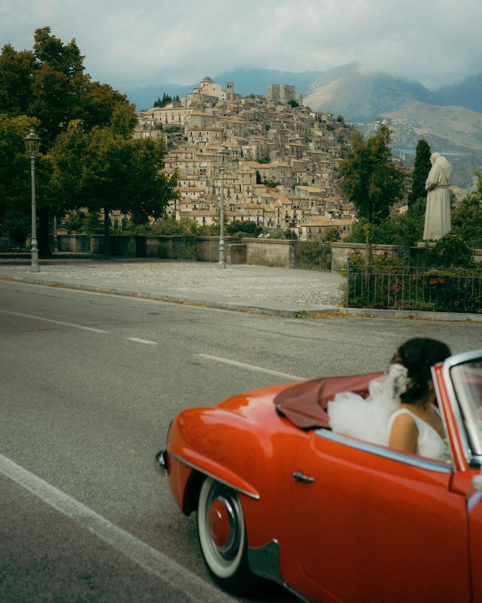 A photo of a bride in a red vintafe mercedes driving past an italian postcard town