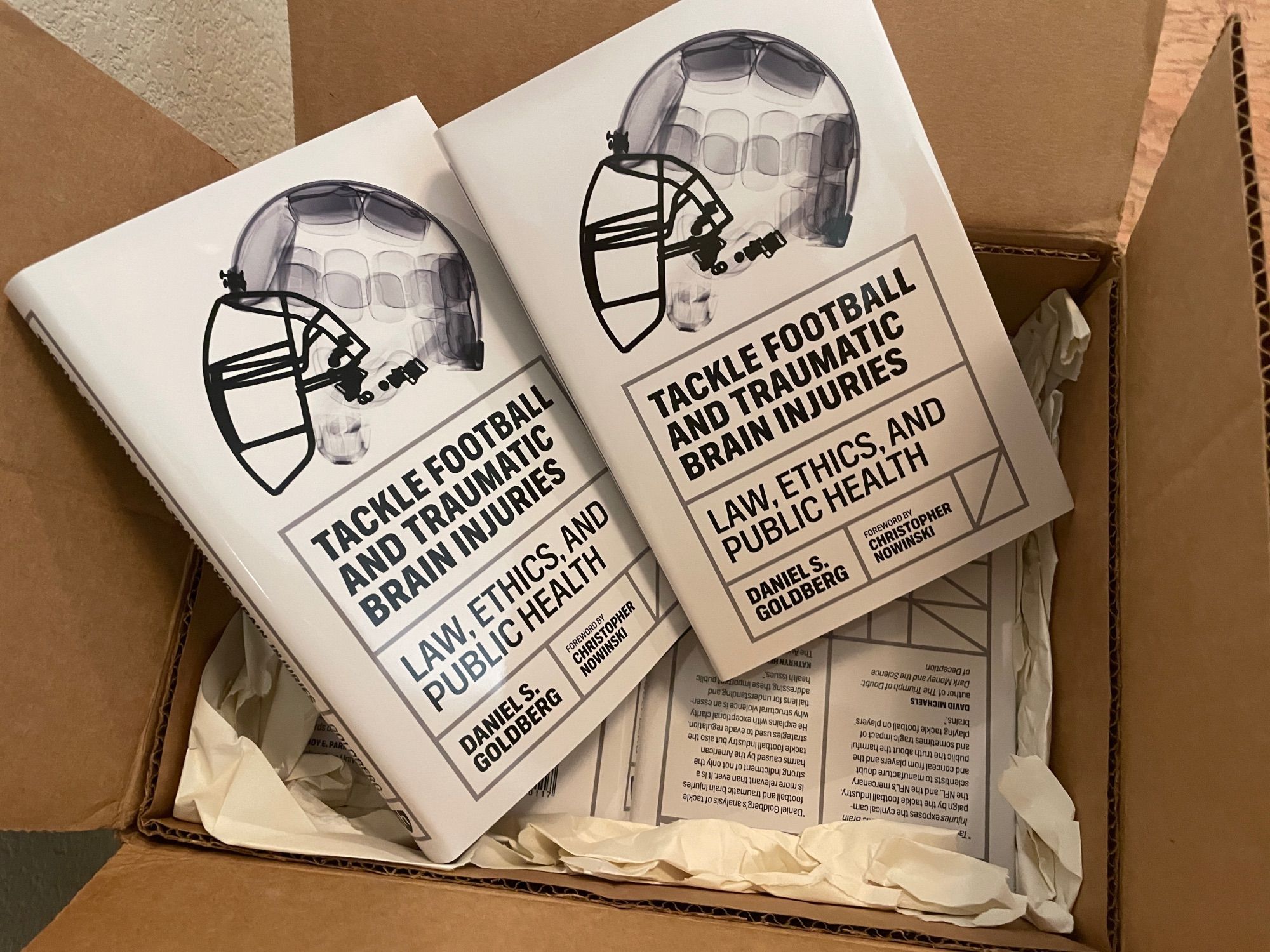 copies of the same book standing up in an open box with more copies lying flat underneath. The title of the book is "Tackle Football and Traumatic Brain Injuries: Law, Ethics, and Public Health" by Daniel S. Goldberg