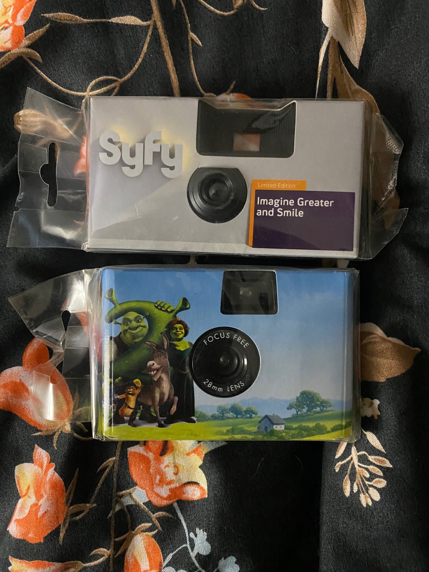 Two disposable 35mm cameras sit atop each other against a cloaca and orange floral blanket. They are each promotional/novelty cameras. 

The top camera is grey with the SyFy channel logo.  

The lower camera is wrapped with a Shrek 2 promotional image, which includes Shrek, Fiona, Donkey, and Puss In Boots standing next to a large, green number 2.