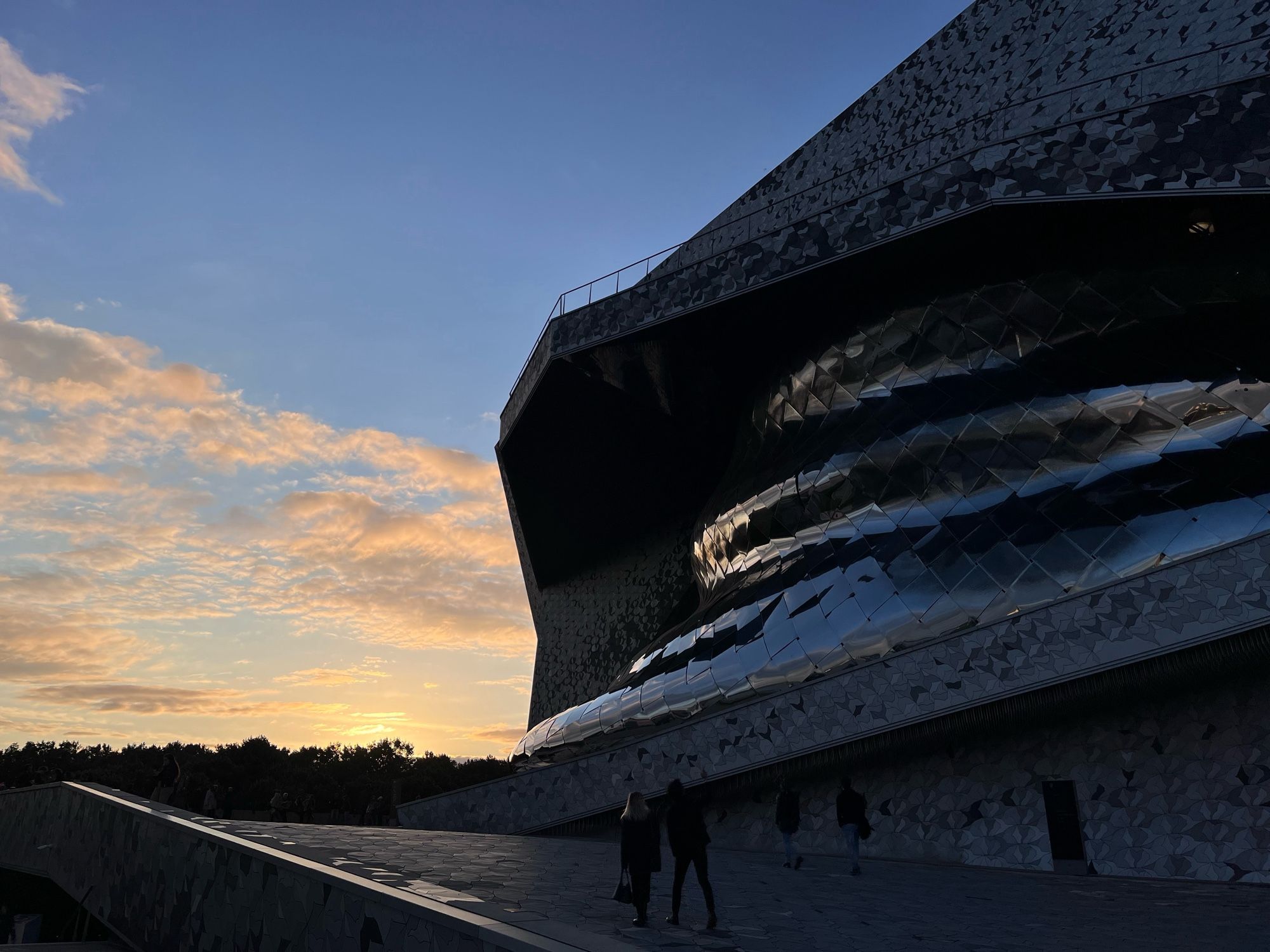 La Philharmonie de Paris (extérieur, façade en contrejour, le soleil se couche derrière les arbres du Parc de la Villette)