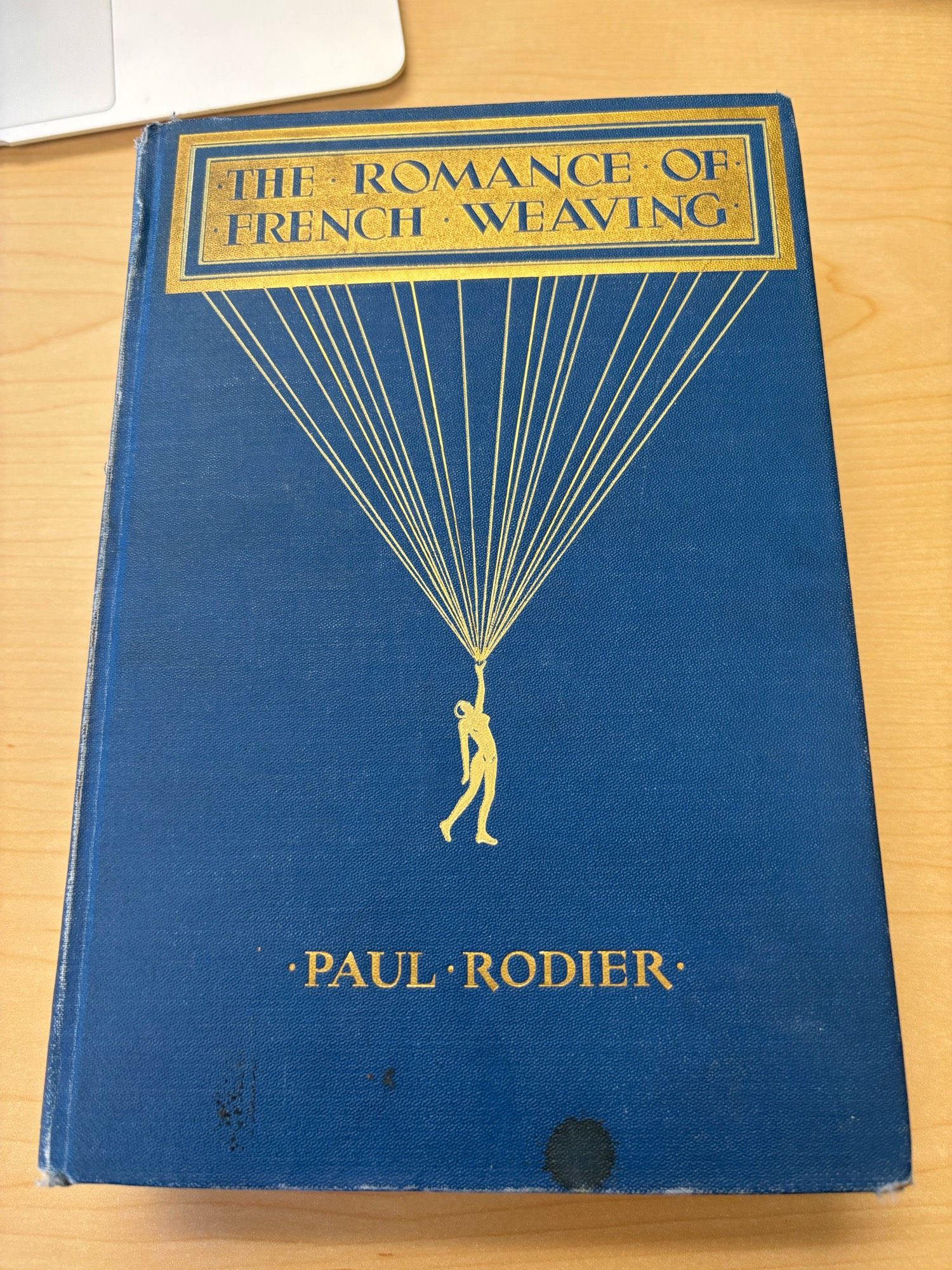 A blue hardcover book with gold writing that says The Romance of French Weaving, Paul Rodier with an image of a (nude?) woman holding a giant handful of warp threads.