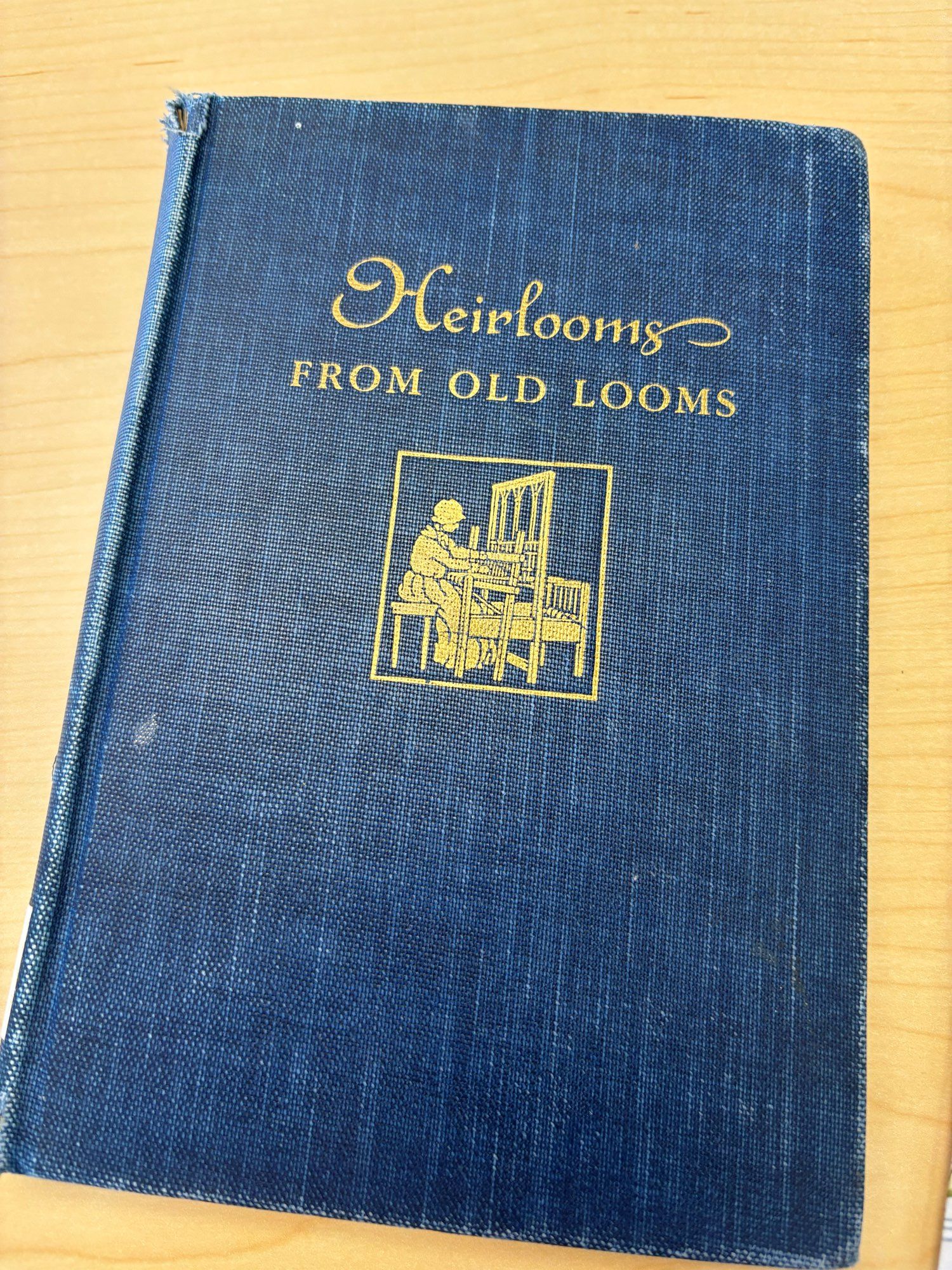 A blue hardcover book with gold writing that says Heirlooms from Old Looms and has a drawing of a 19th or 18th century woman weaving at a large loom.