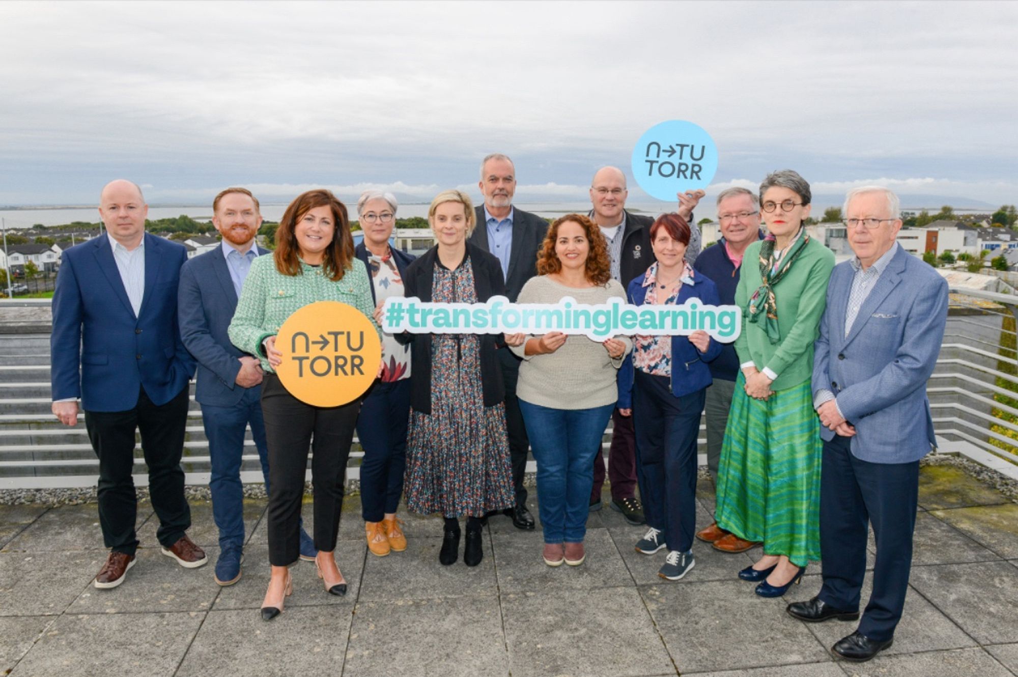 A group of people lined up, outside on a balcony with a view of Galway Bay. They are holding up signs with N-TUTORR branding and the #transforminglearning letters.