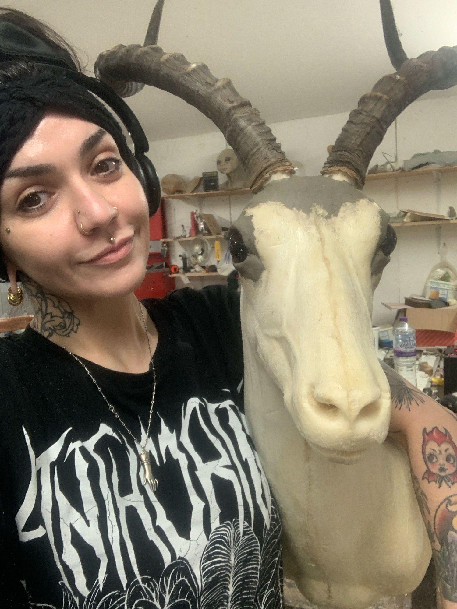 Me with headphones and knitted headband wearing a black metal inspired plant tshirt that says ‘anthurium’ on it, standing with my arm draped around the neck of a foam impala form that I was prepping for taxidermy. I’d already fixed the horns in place & set the eyes with clay.