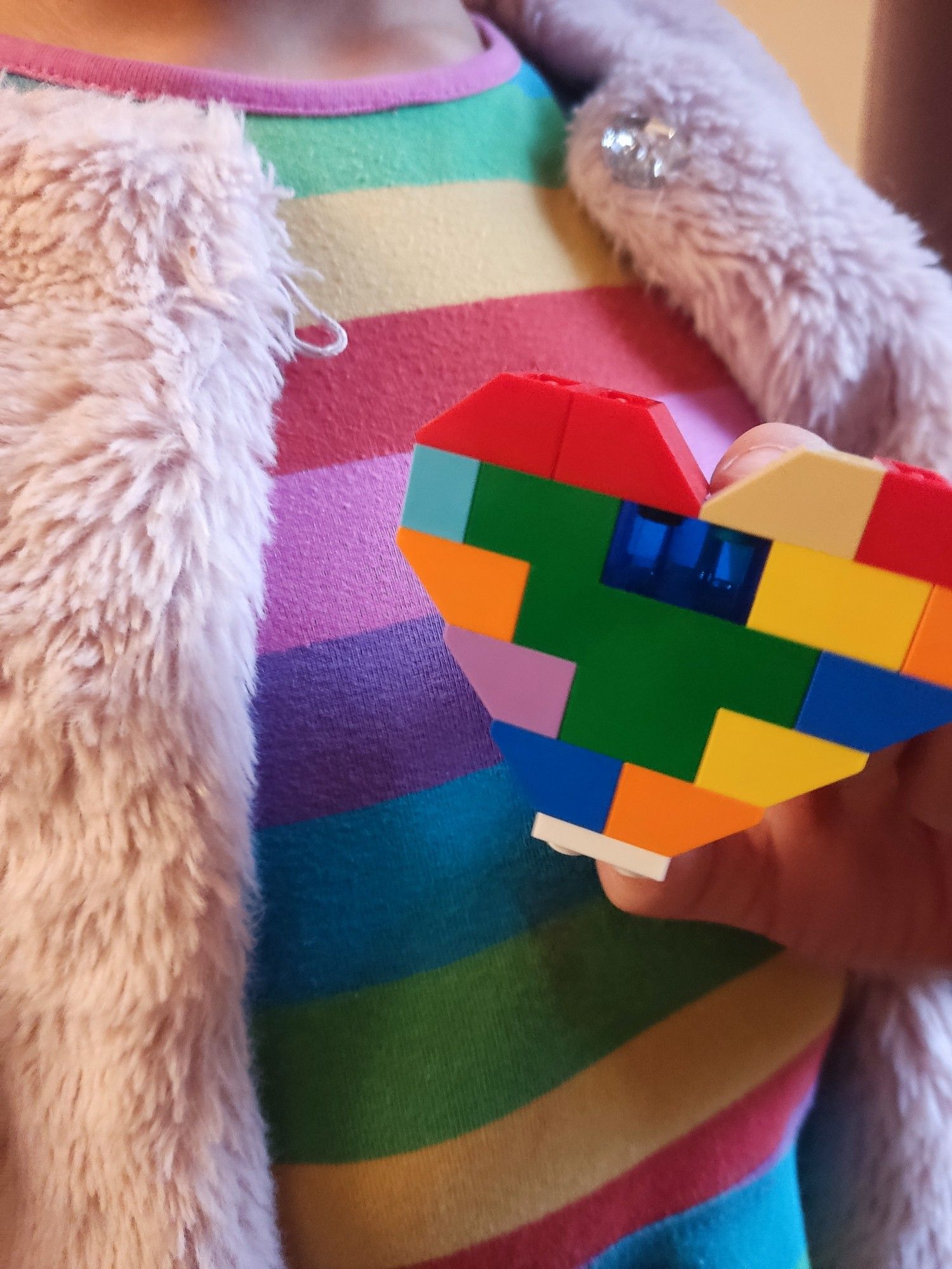 Kind in Regenbogen-Kleid und rosa Fell-Jacke hält Regenbogen-Herz aus Lego. Bildausschnitt auf Brustkorbhöhe