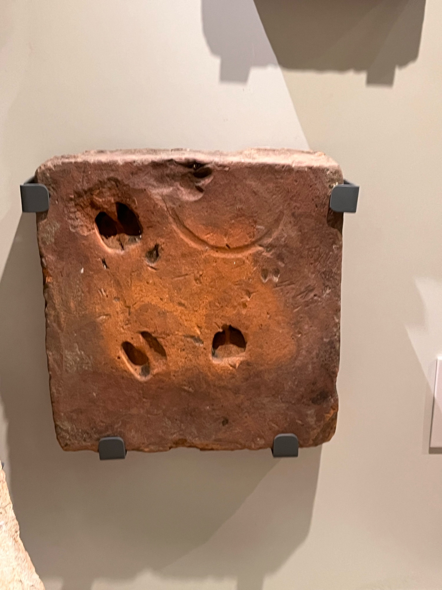 A wall-mounted square Romano-Briton terracotta tile on display in a museum case. It is a deep umber and has six different imprints: three small deer hoofs, two dog paws, and the edge of a sandal.