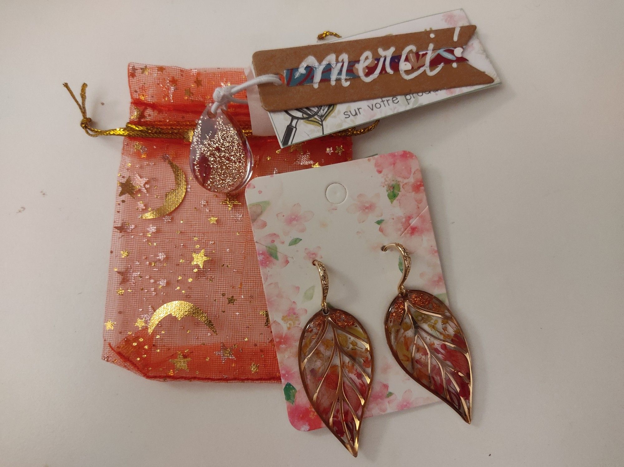 Des boucles d'oreilles en forme de feuille, sur une pochette en tissu rouge avec des étoiles, et un petit carton de remerciement