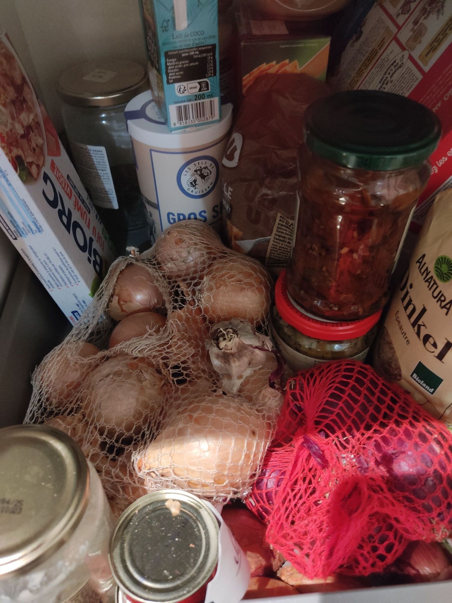 L'intérieur d'un placard avec des filets d'oignons rouges et jaunes, quelques échalotes et un peu d'ail. On voit d'autres paquets/bocaux (tofu fumé, gros sel, lait de coco, épeautre, lasagnes, concentré de tomate, tahini...)