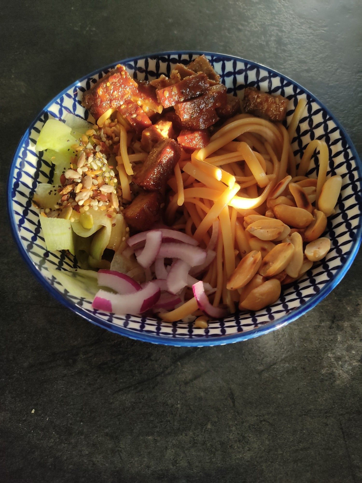 Un bol chinois éclairé par un rayon de soleil avec de l'oignon rouge émincé, des lanières de concombre parsemées de graines et d'oignons frits, des dés de tofu caramélisé, des linguine (dans une sauce au soja, nuoc mam et miel) avec des cacahuètes torréfiées par dessus