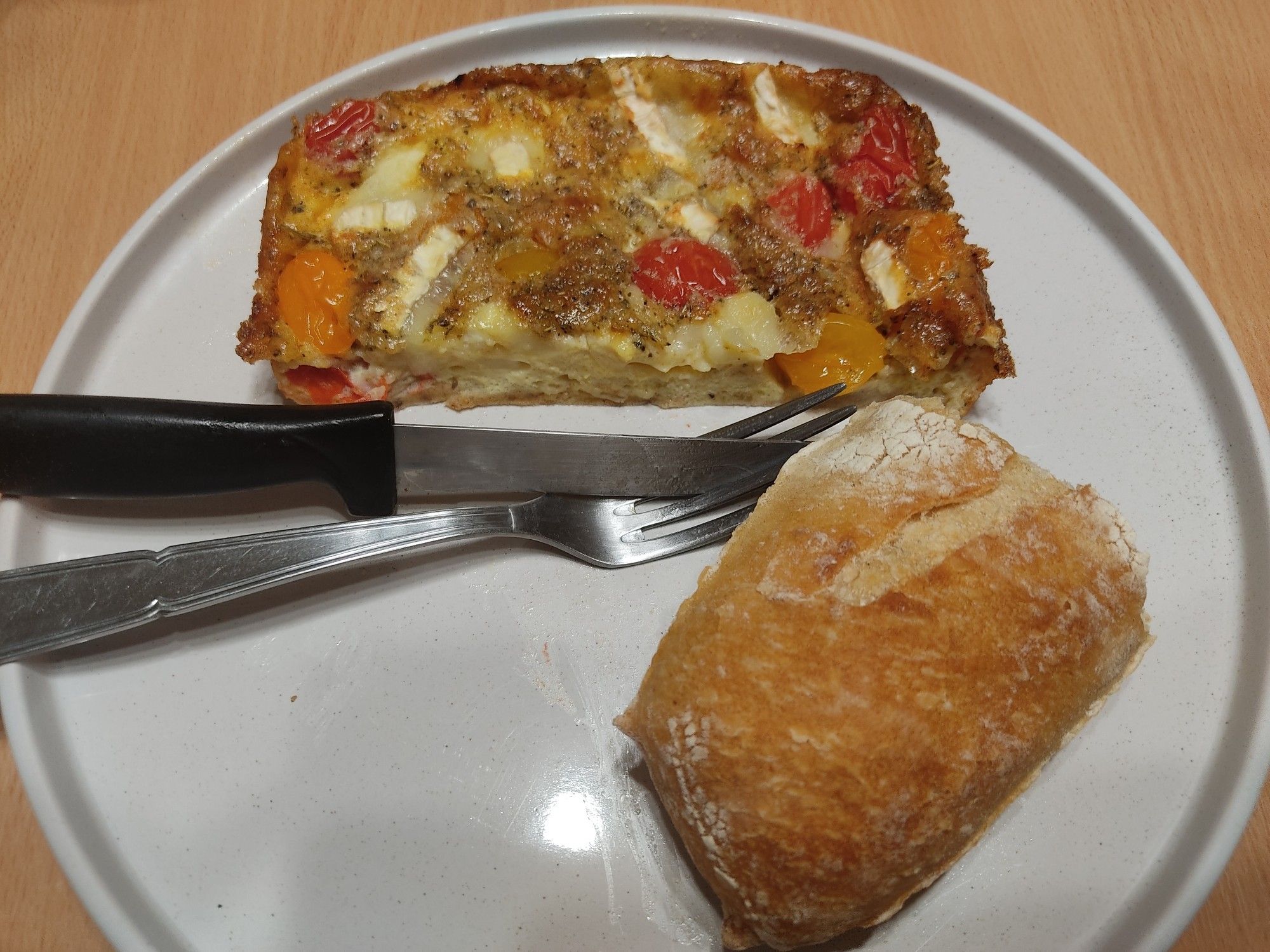 Une assiette couleur crème avec un couteau à steak et une fourchette, en haut le fameux clafoutiti aux tomates cerises et au chèvre et un morceau de baguette en bas de l'assiette