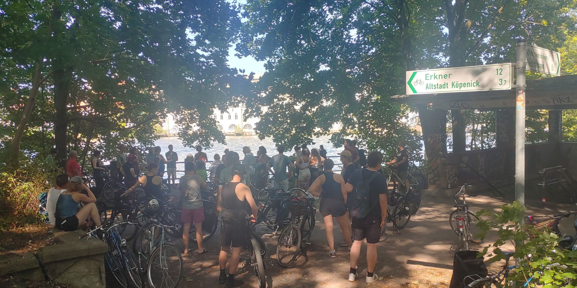 Eine große Gruppe Radfahrender hört einem Guide zu. Sie stehen unter schattigen Bäumen an der Spree. Rechts im Bild ein Radfahrschild nach Erkner und zur Altstadt Köpenick.