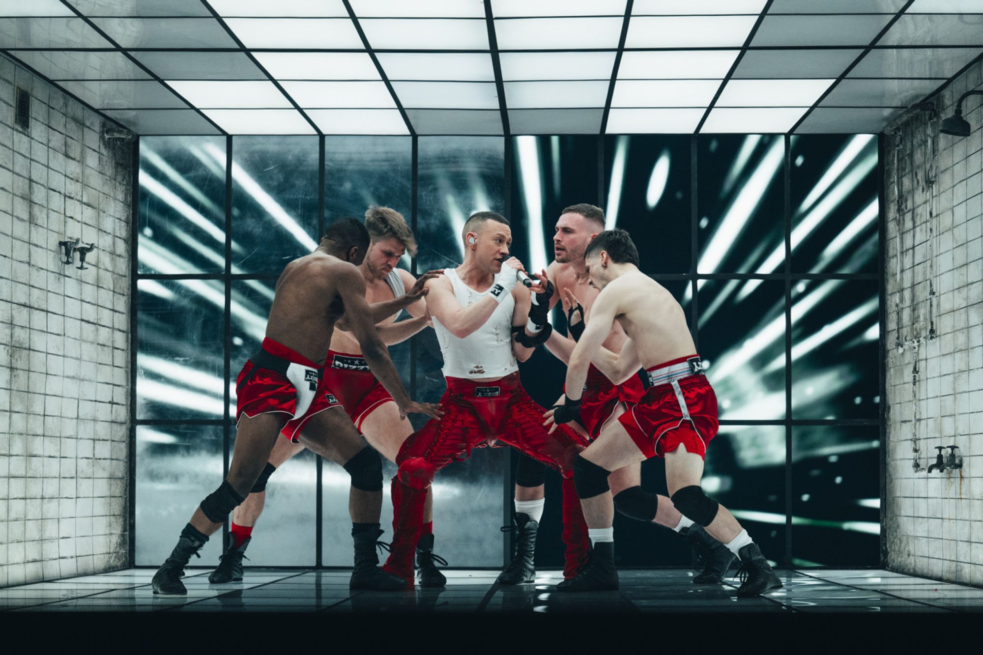 Olly Alexander (representing Great Britain at Eurovision), rehearsing his song. He's in a set designed to look like a dingy gym shower room. He's wearing a white singlet and leather pants, surrounded by four shirtless men wearing red athletic shorts. One has what appears to be some kind of groin protector over the top of his shorts.